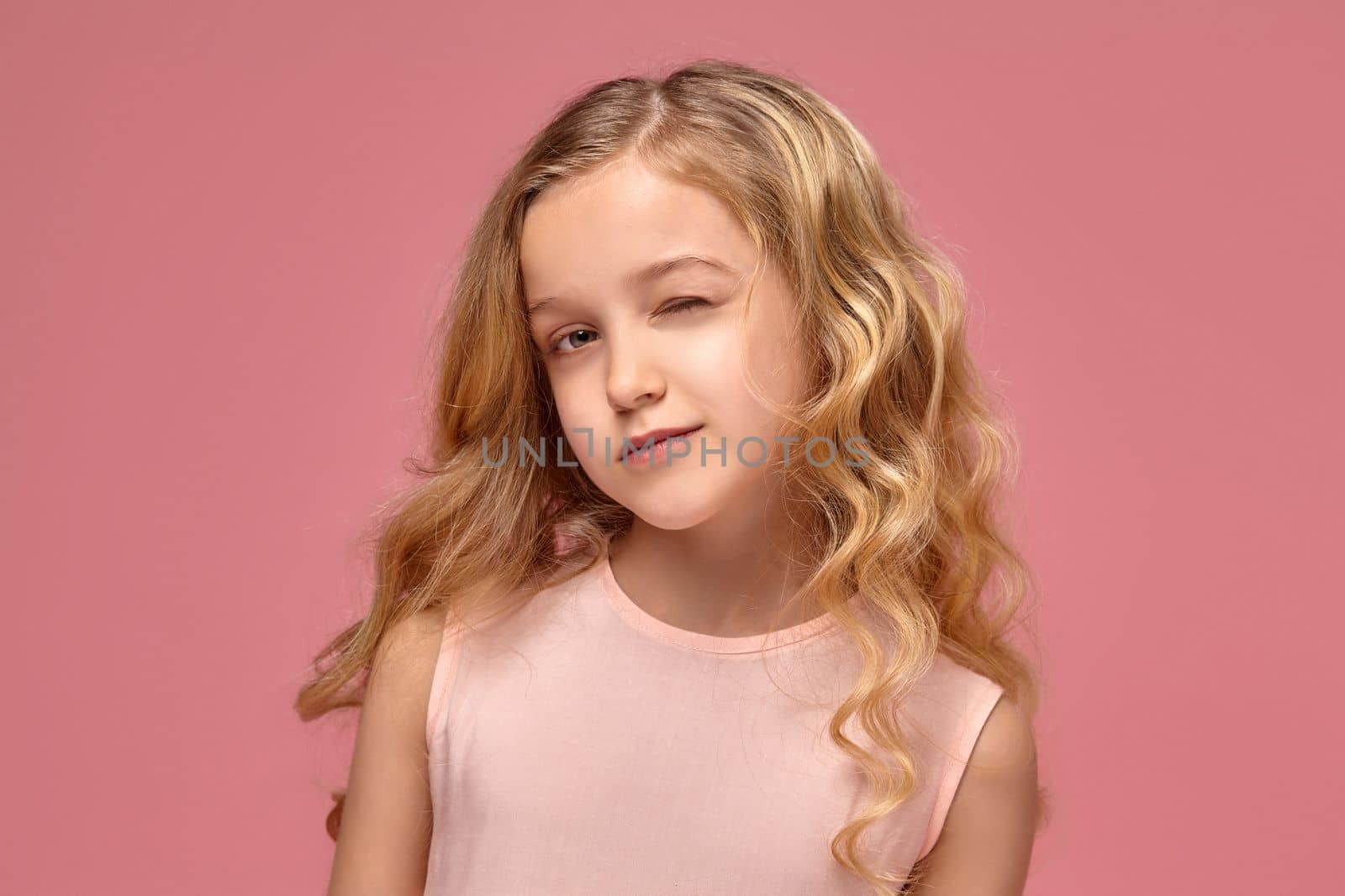 Beautiful little girl with a blond curly hair, in a pink dress winks into the camera, on a pink background