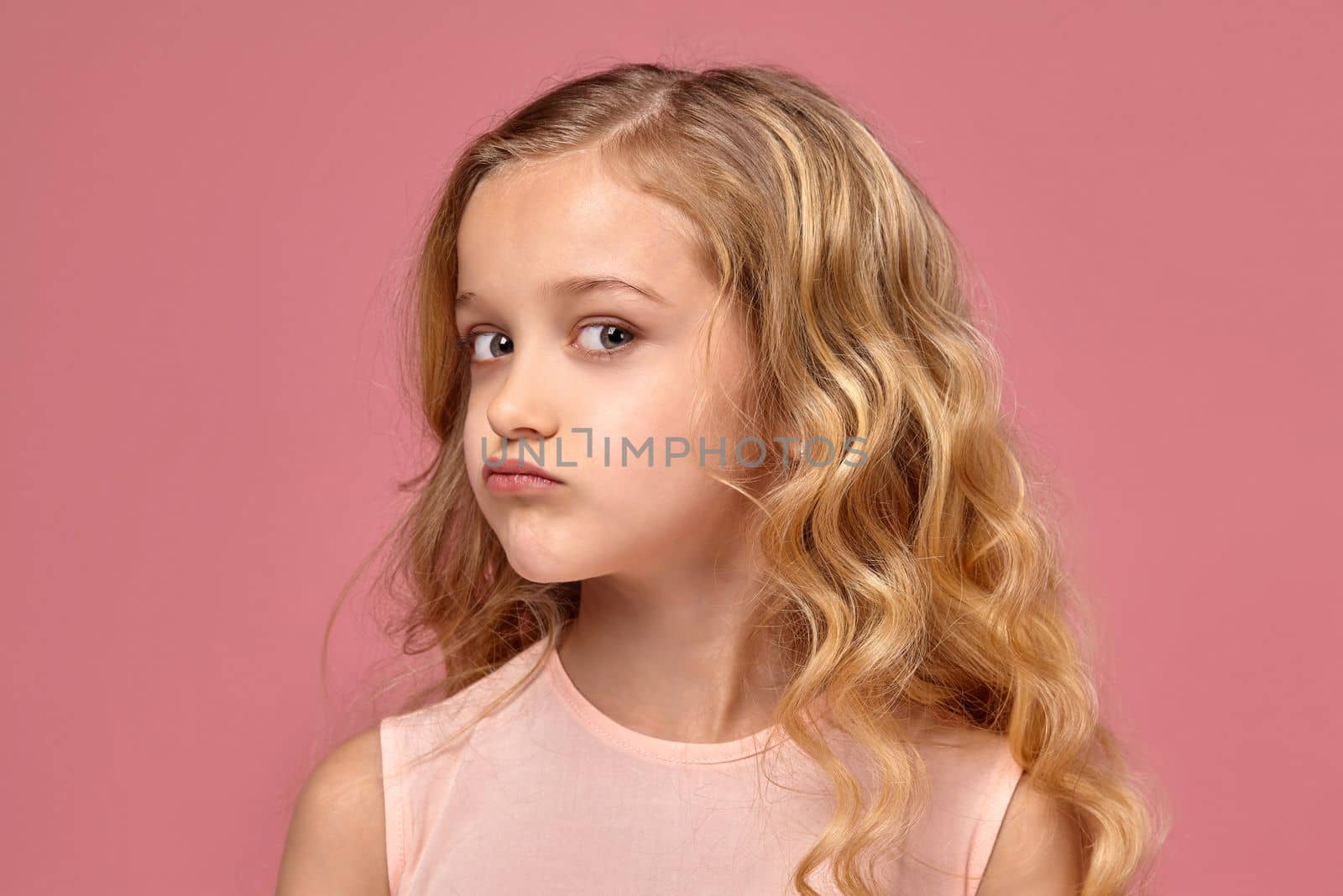 Beautiful little girl with a blond curly hair, in a pink dress poses for the camera and looks offended, on a pink background