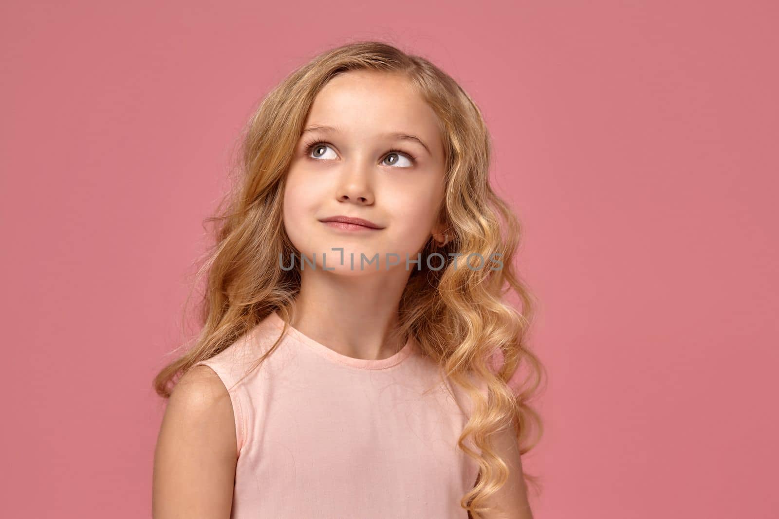 Little girl with a blond curly hair, in a pink dress is looking away, on a pink background