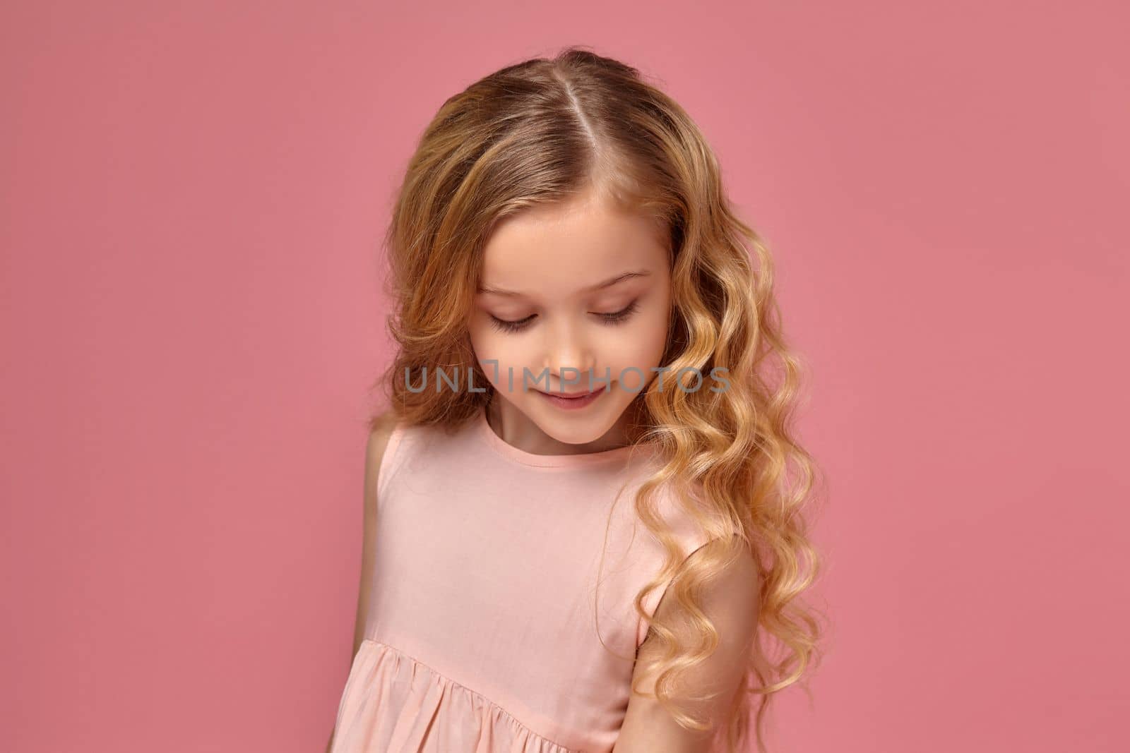 Pretty little girl with a blond curly hair, in a pink dress is posing fot the camera and looking shy, on a pink background