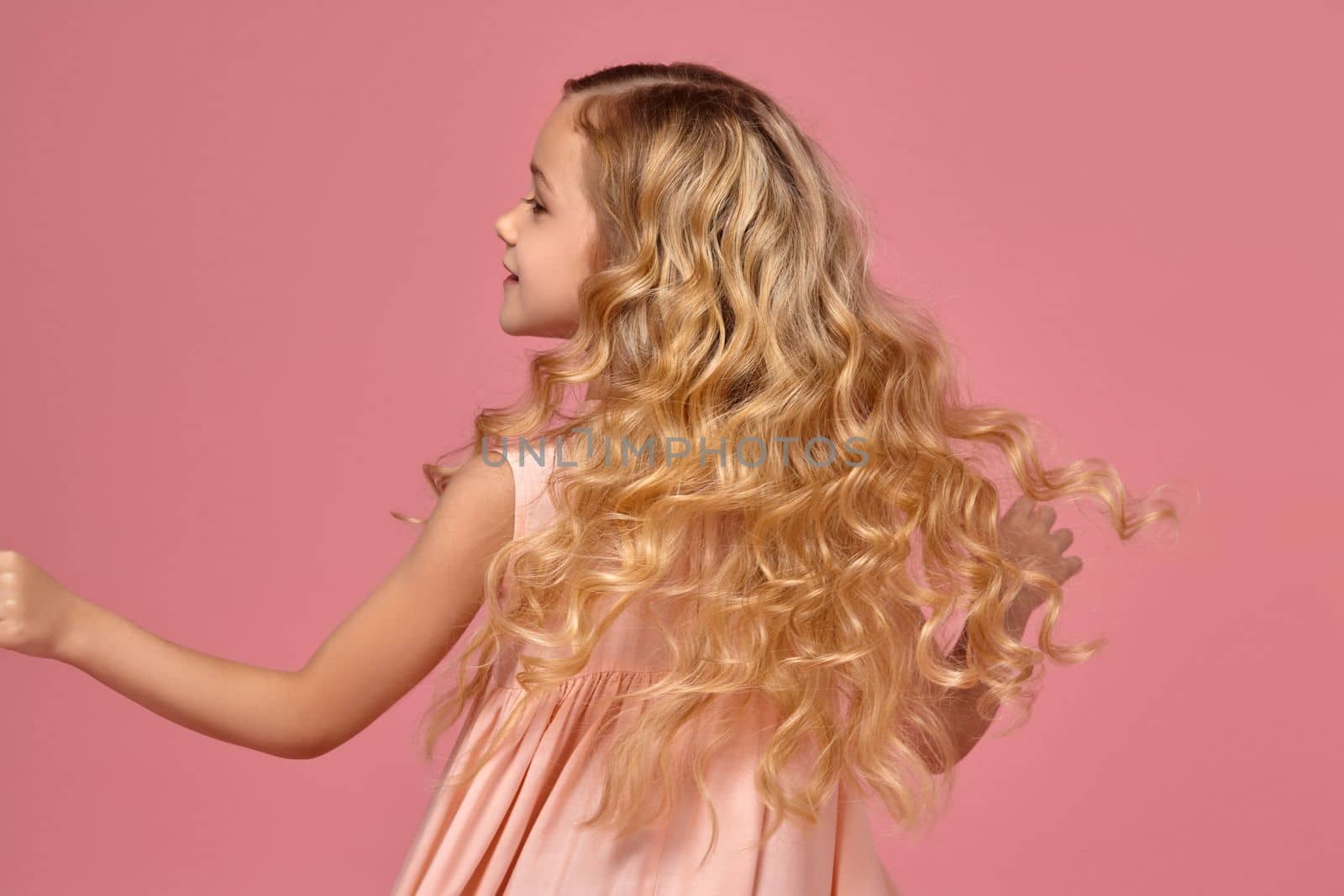 Beautiful little girl with a blond curly hair, in a pink dress is spinning around fot the camera, on a pink background