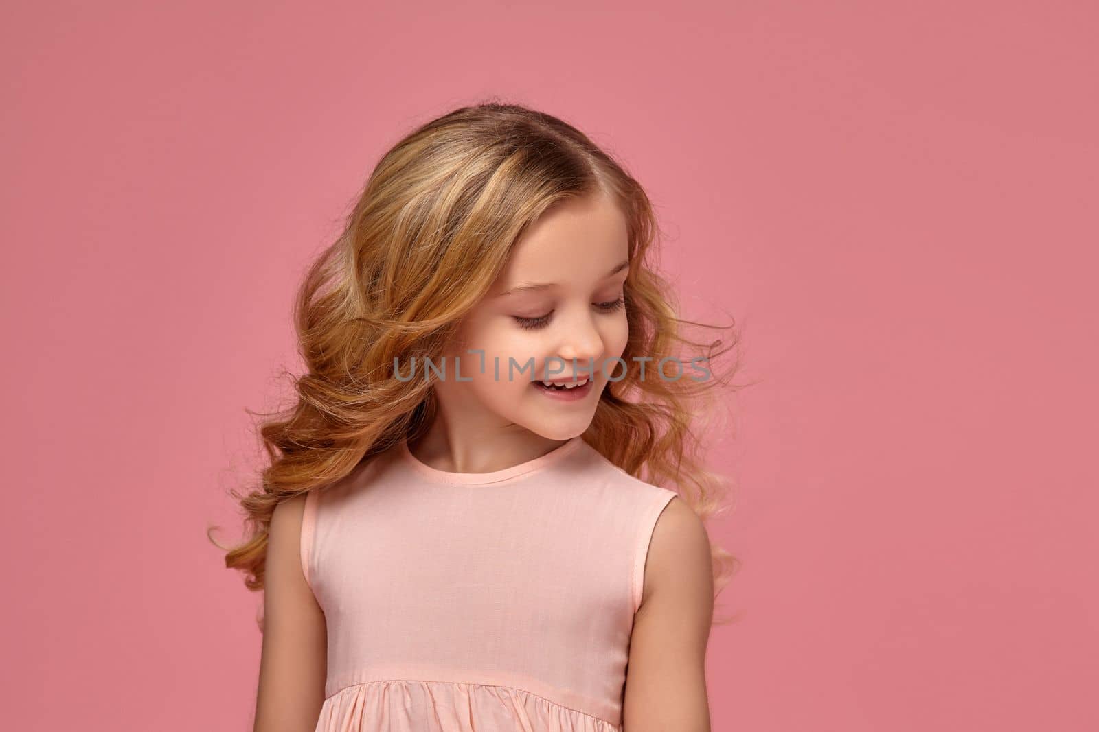 Beautiful little girl with a blond curly hair, in a pink dress is posing for the camera and looking down, on a pink background
