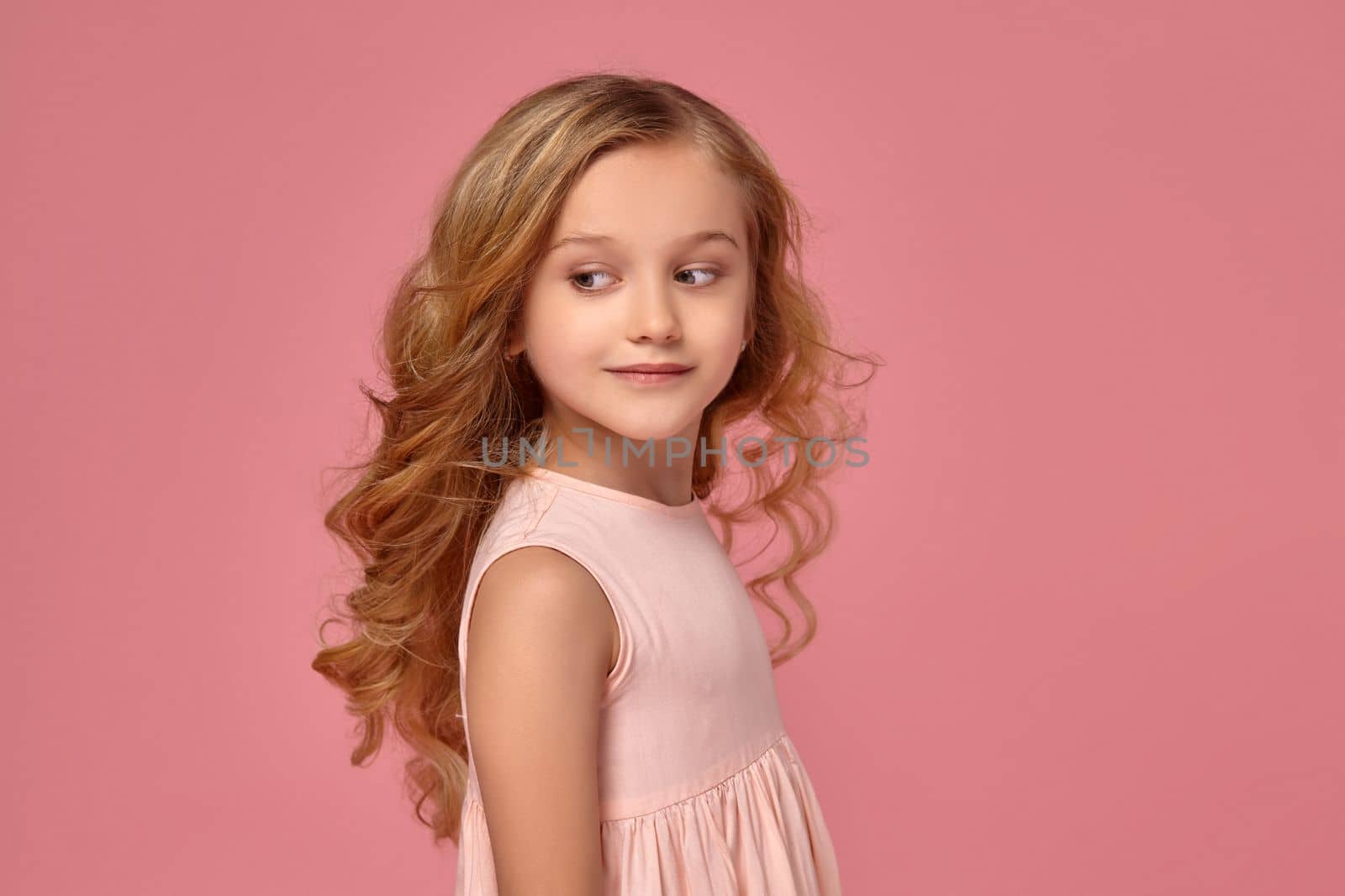 Amazing little girl with a blond curly hair, in a pink dress is posing for the camera and looking down, on a pink background