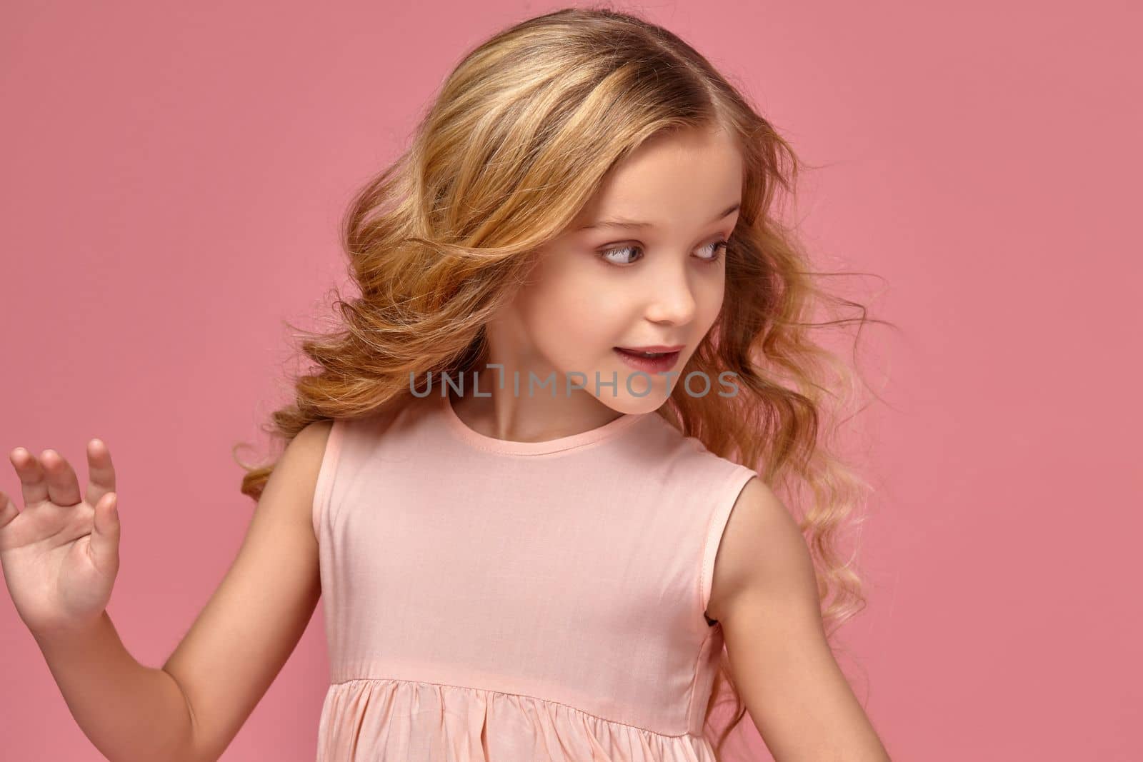 Funny little girl with a blond curly hair, in a pink dress is posing for the camera and looking away, on a pink background