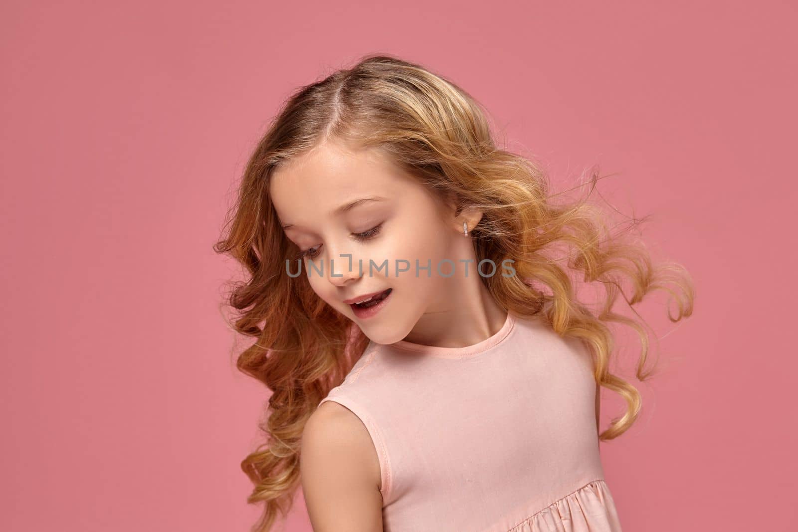 Little girl with a blond curly hair, in a pink dress poses for the camera and looks back, on a pink background
