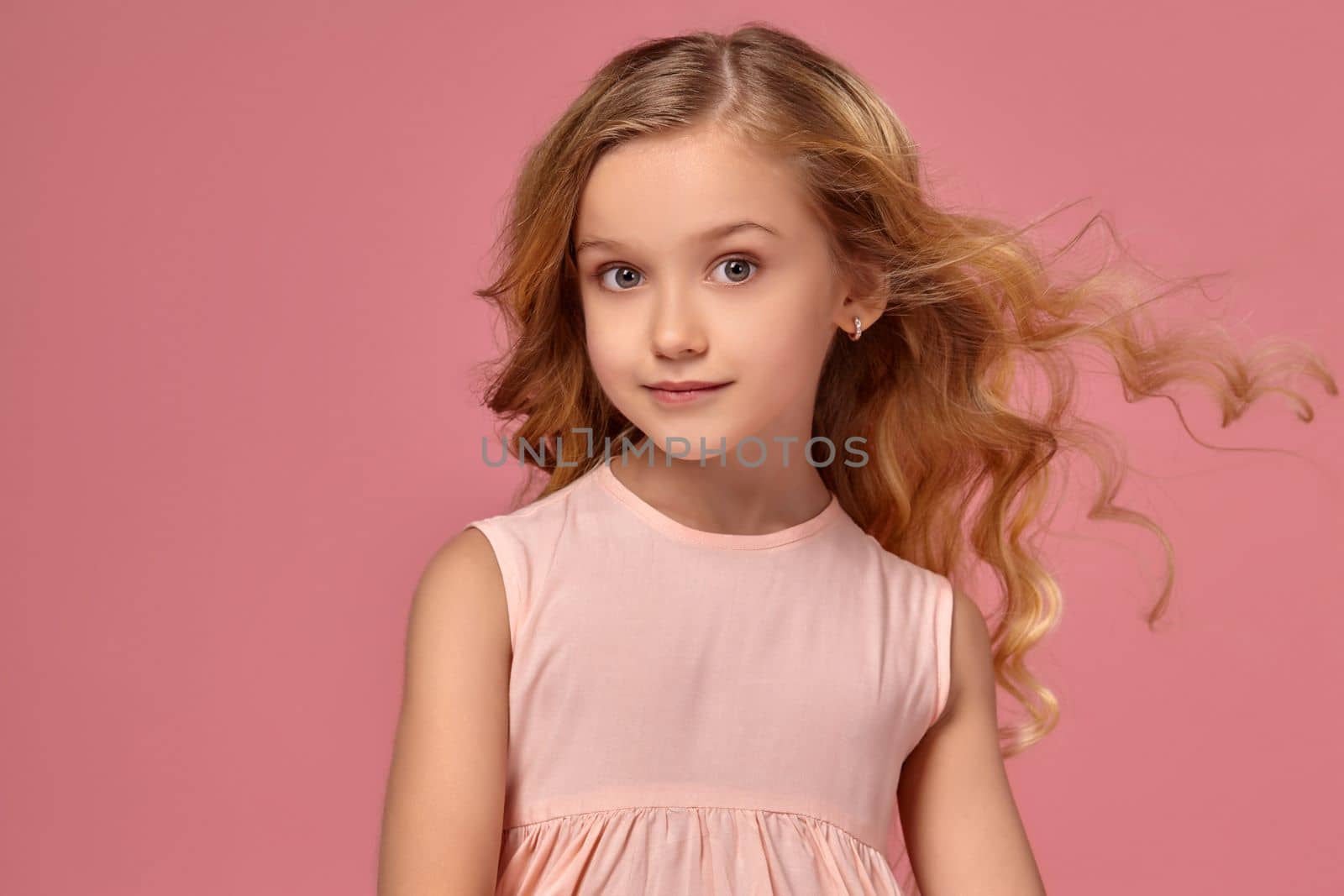 Beautiful little girl with a blond curly hair, in a pink dress is posing for the camera and her hair is fluttering, on a pink background