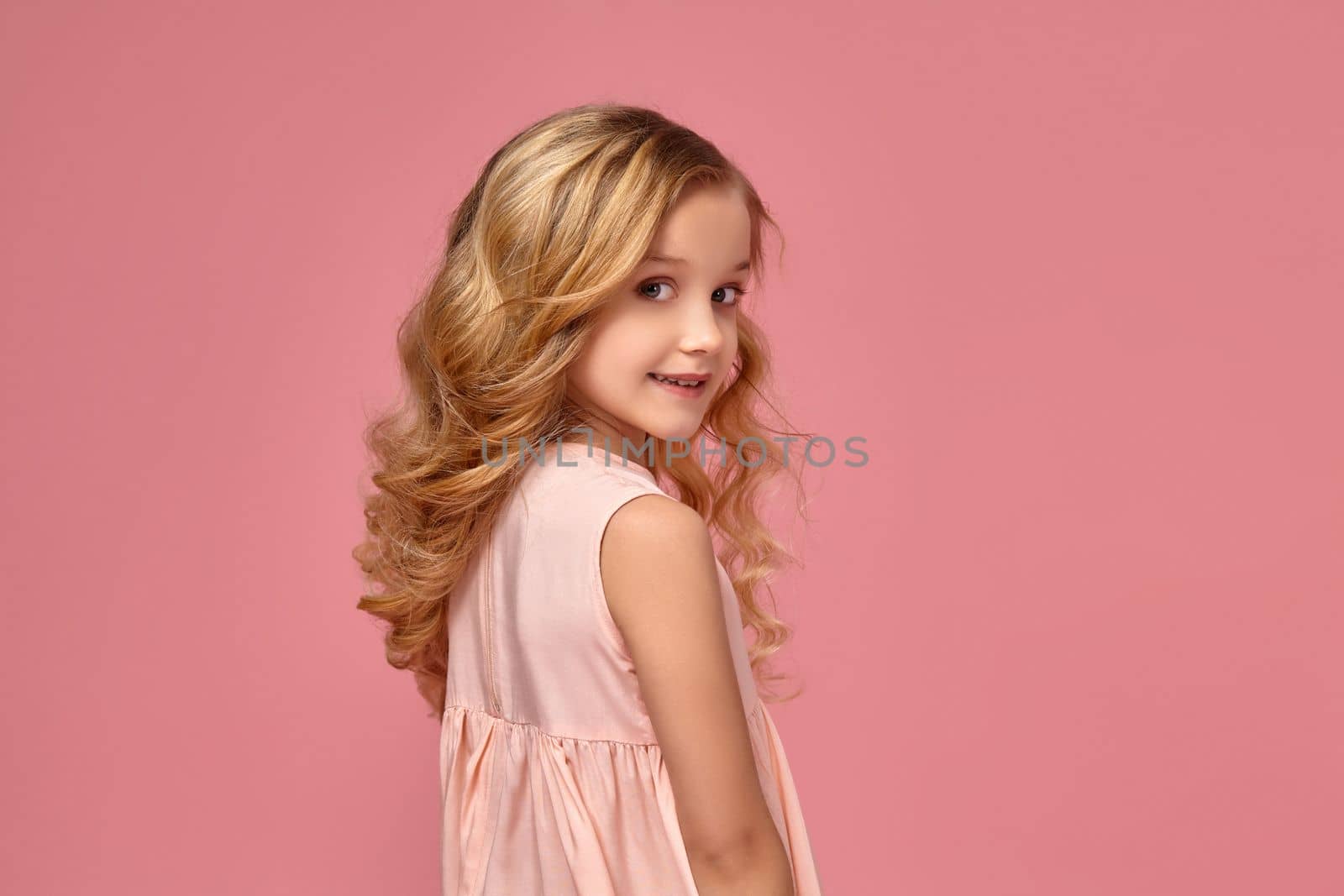 Little girl with a blond curly hair, in a pink dress is posing for the camera by nazarovsergey