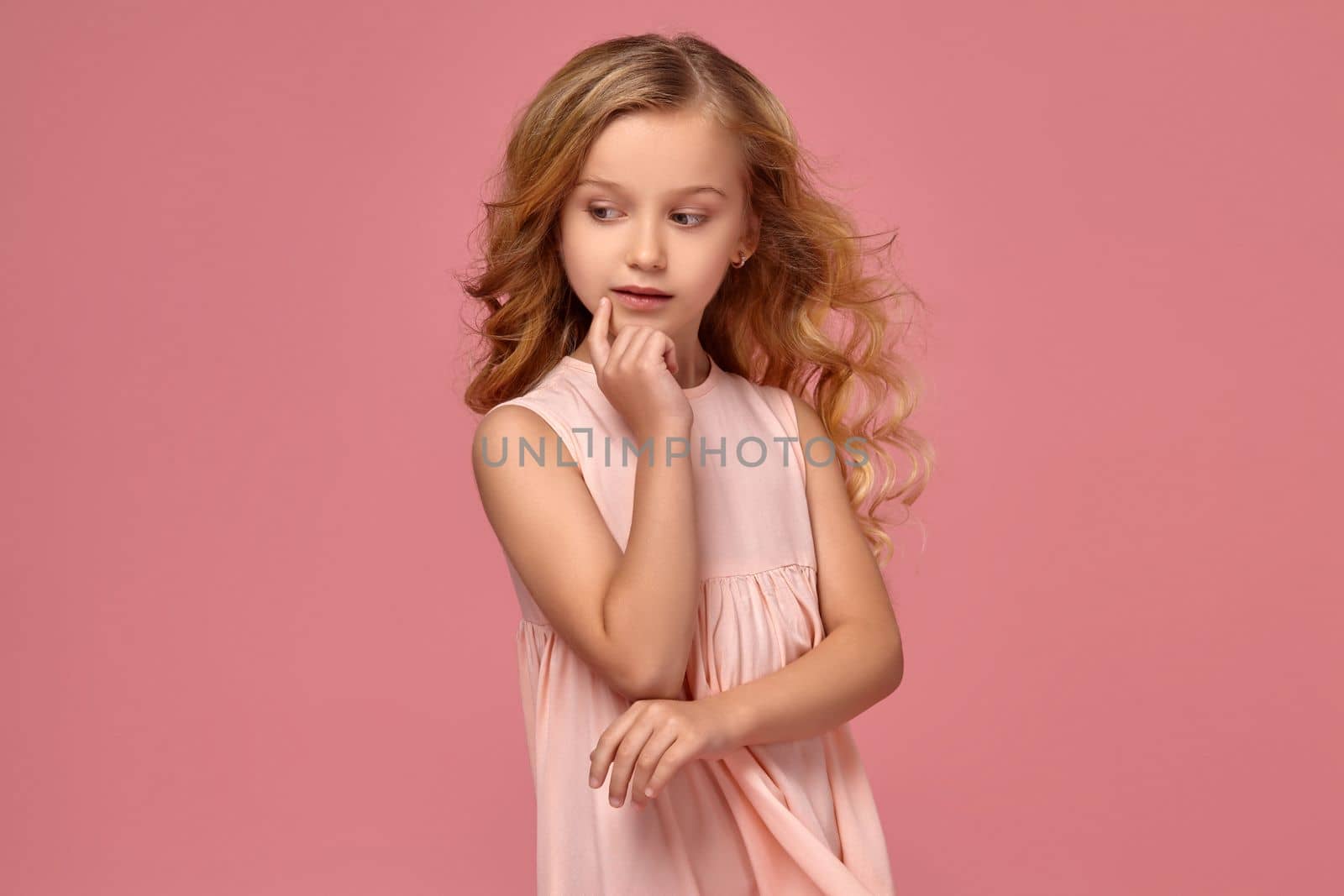 Charming little girl with a blond curly hair, in a pink dress poses for the camera and looks thoughtful, on a pink background