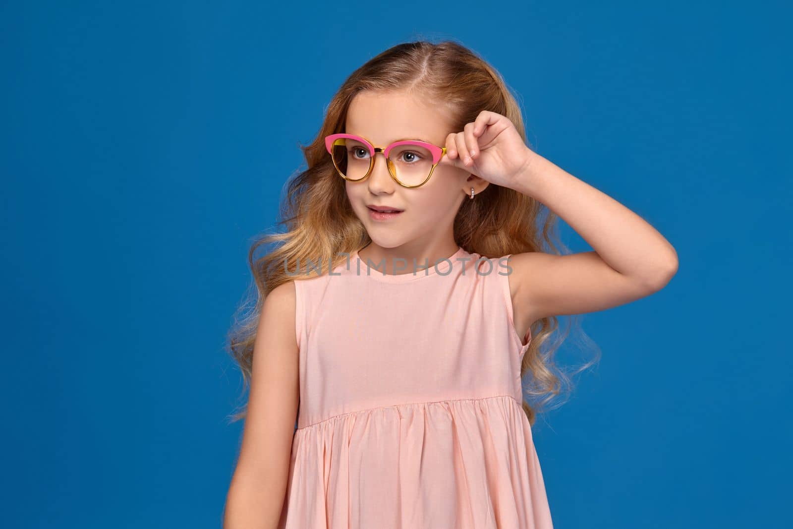 Funny little girl in a pink dress is straightense her fashionable glasses, standing on a blue background.