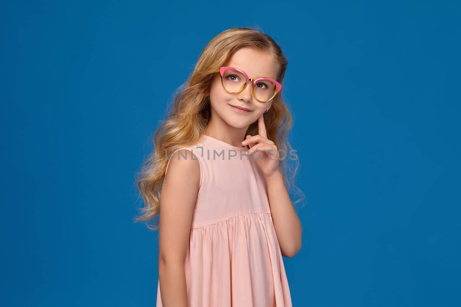 Modern little girl in a pink dress and a fashionable glasses is touching her cheek, standing on a blue background.