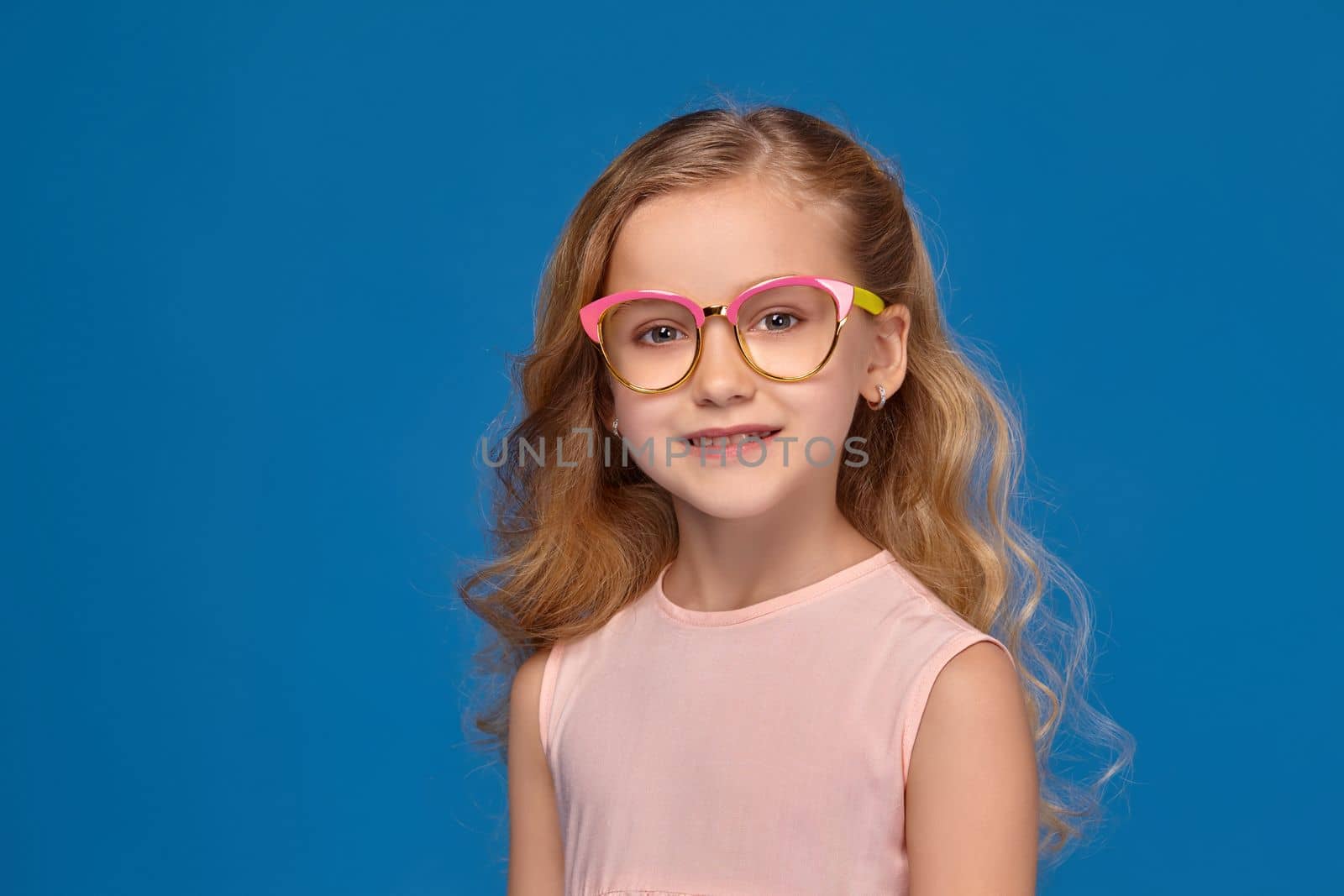 Modern little girl in a pink dress and a fashionable glasses is holding a candy, standing on a blue background.