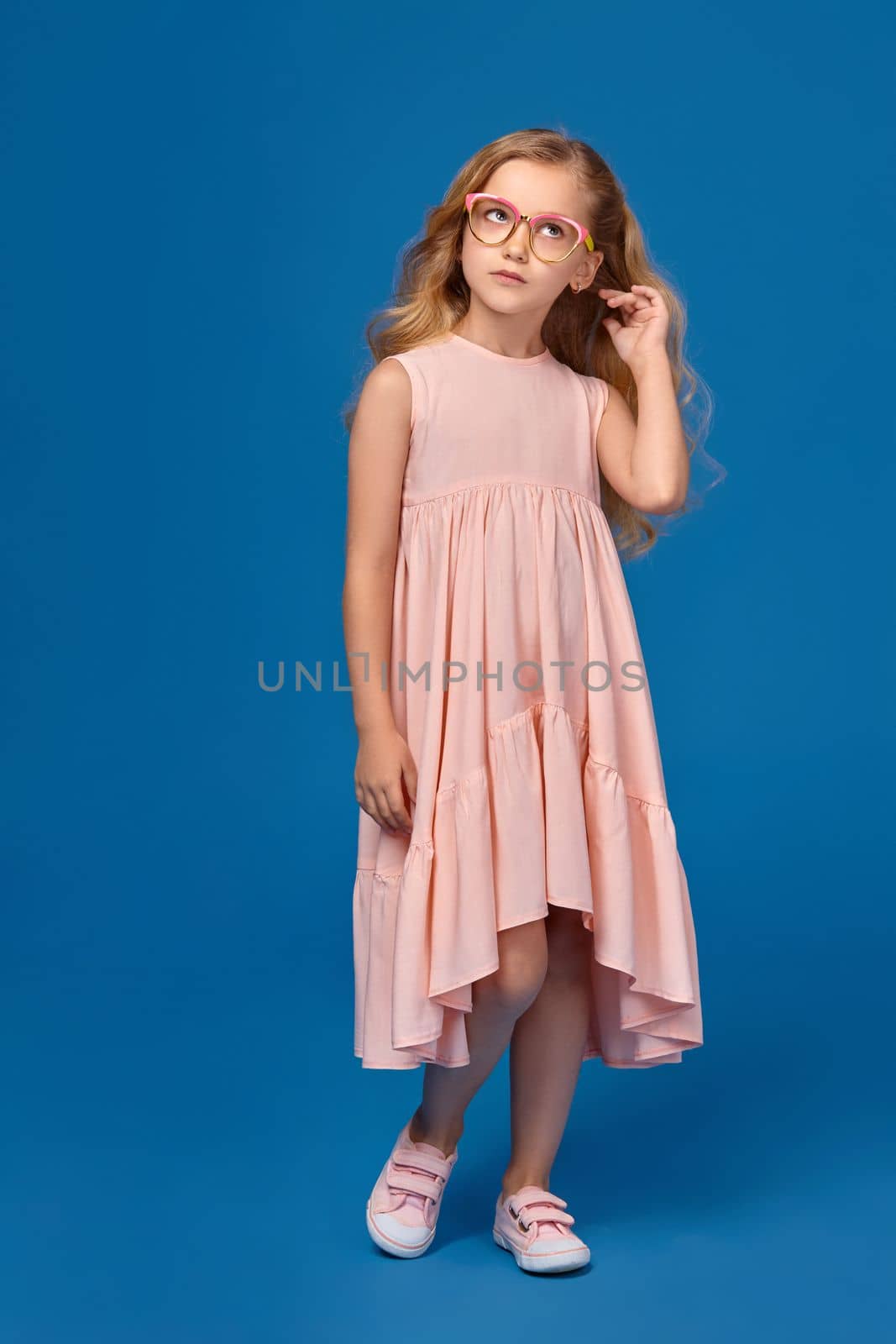 Modern little girl in a pink dress and a fashionable glasses is looking up, standing on a blue background.