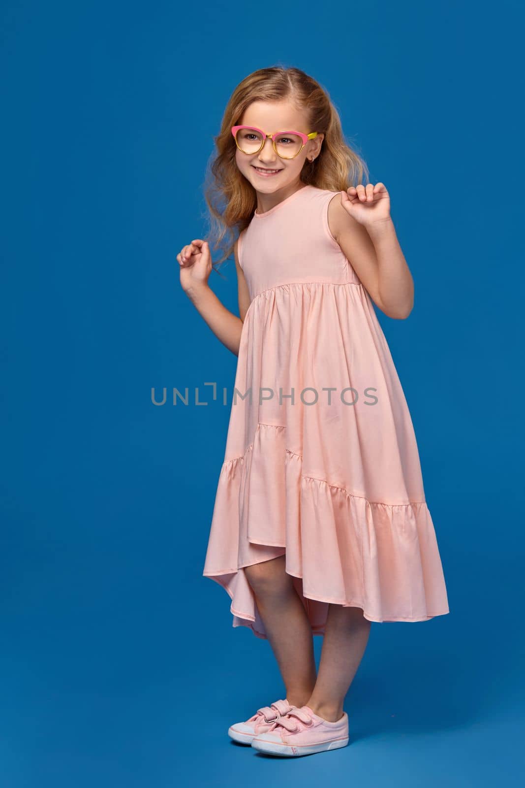 Modern little girl in a pink dress, fashionable glasses and a pink sneakers is dancing, on a blue background.