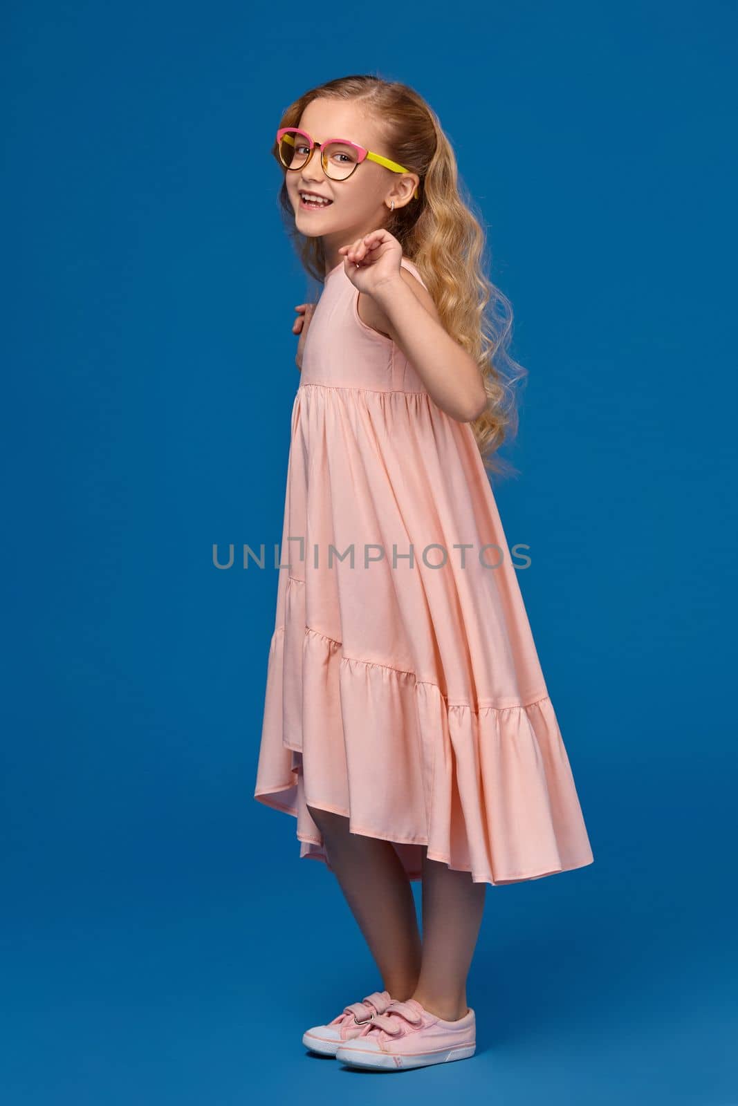 beautiful little girl in a pink dress, fashionable glasses and a pink sneakers is dancing, on a blue background.