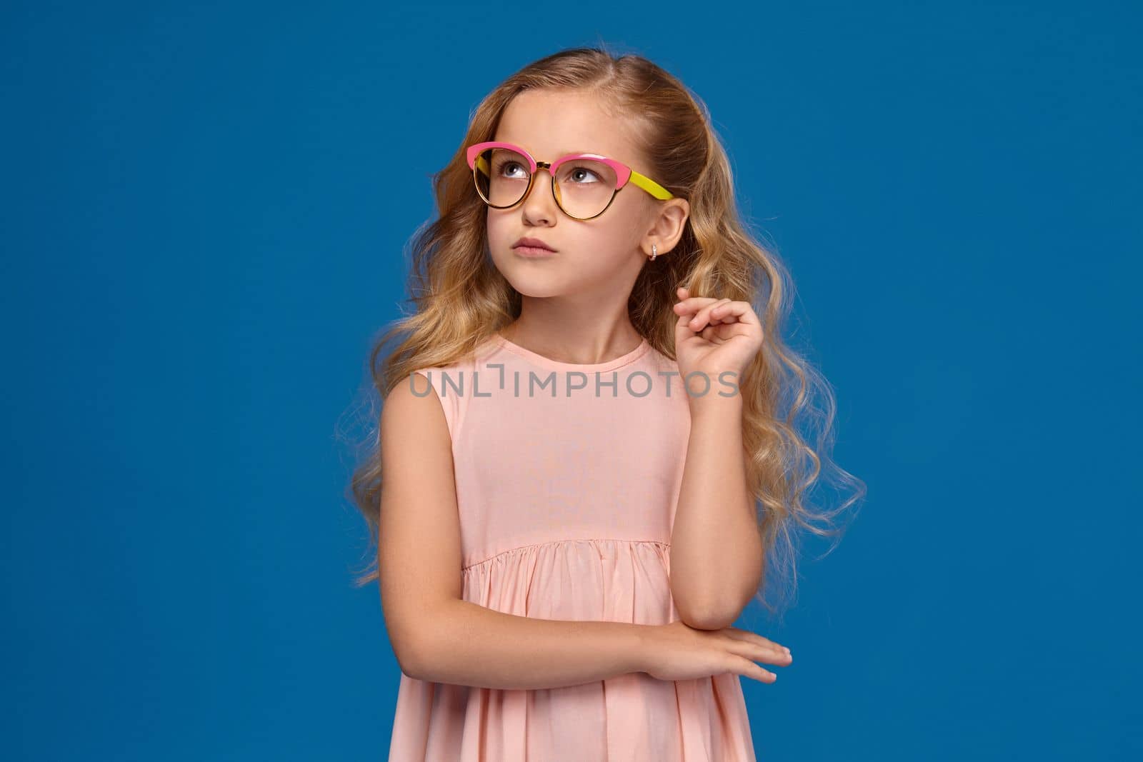 Pretty little girl in a pink dress and a fashionable glasses is looking up thoughtfully, standing on a blue background.