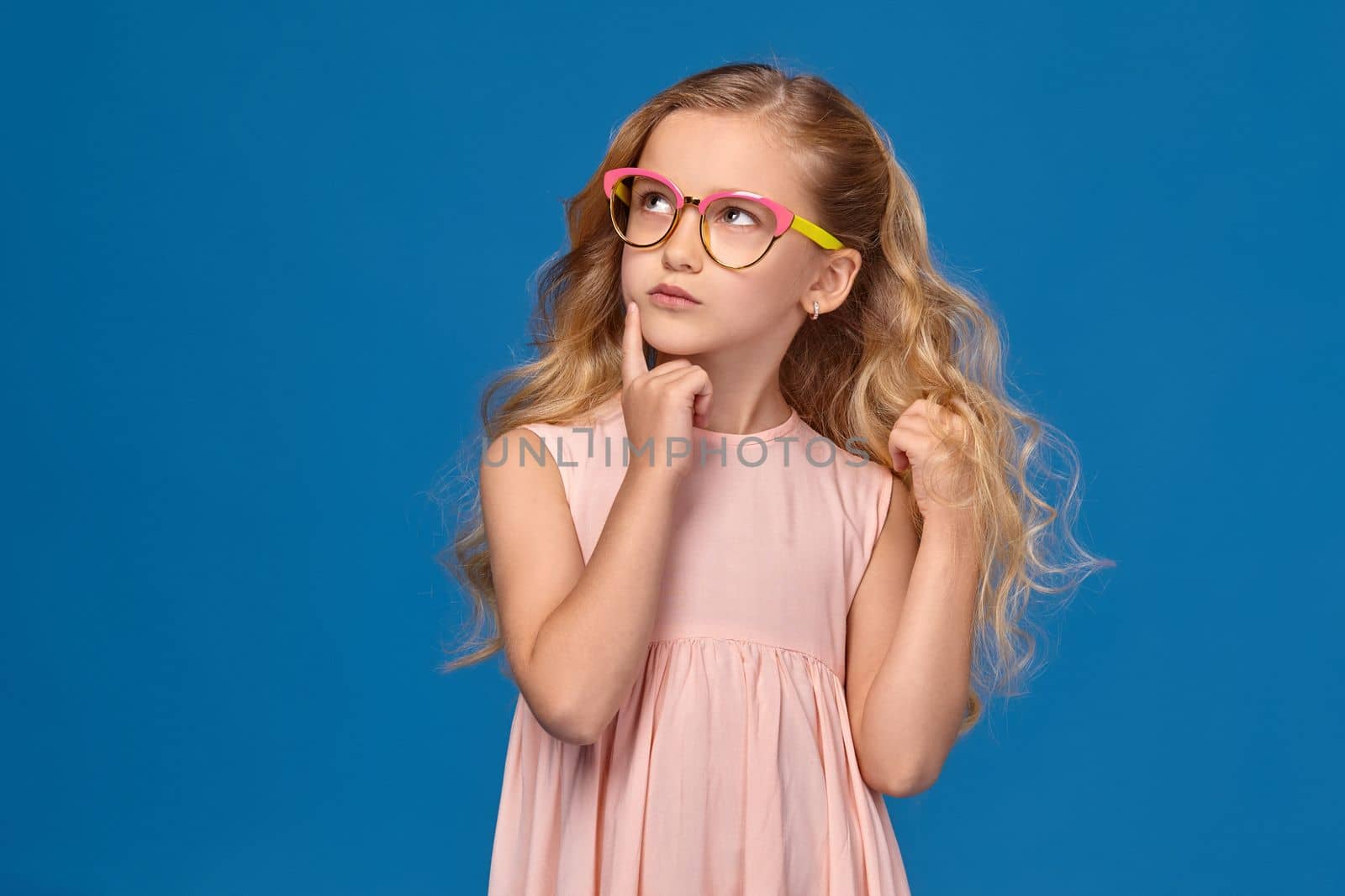 Beautiful little girl in a pink dress and a fashionable glasses is looking up thoughtfully, standing on a blue background.