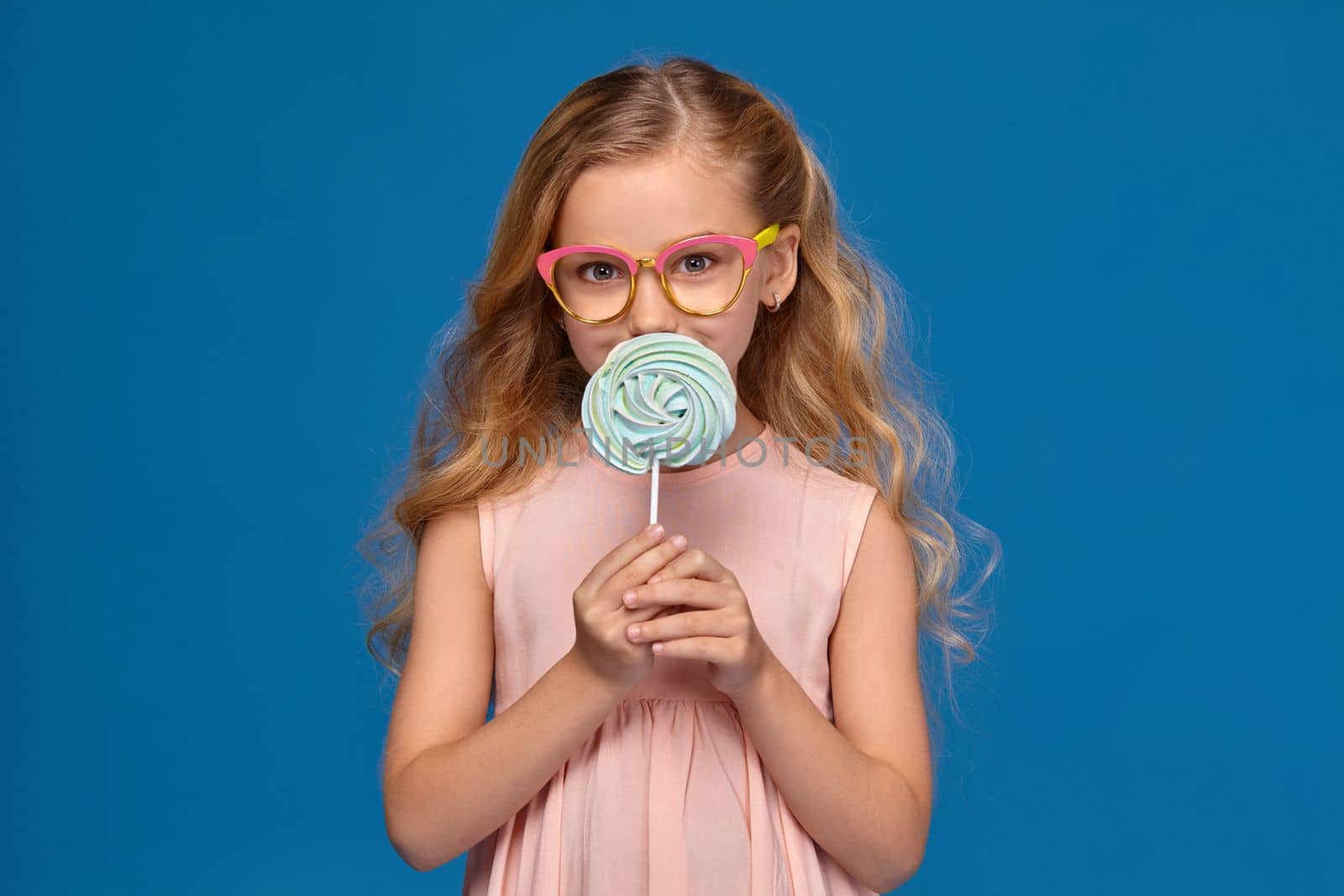 Modern little girl in a pink dress and a fashionable glasses is holding a candy, standing on a blue background.