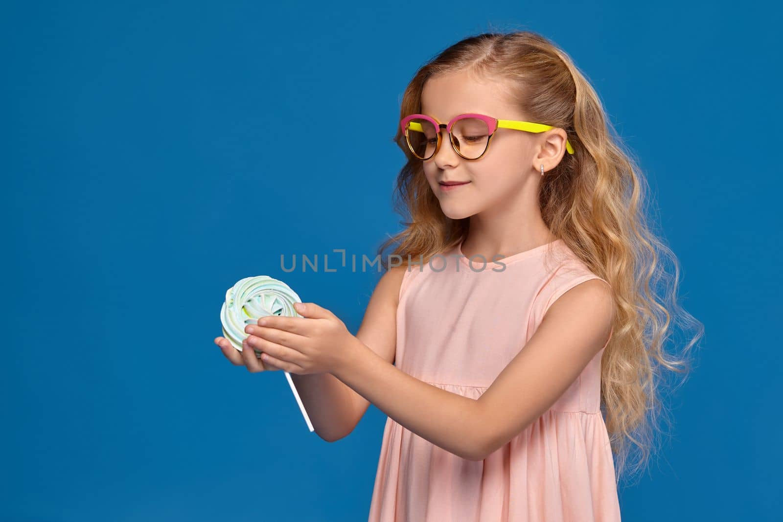 Charming little girl in a pink dress and a fashionable glasses is holding a candy, standing on a blue background.