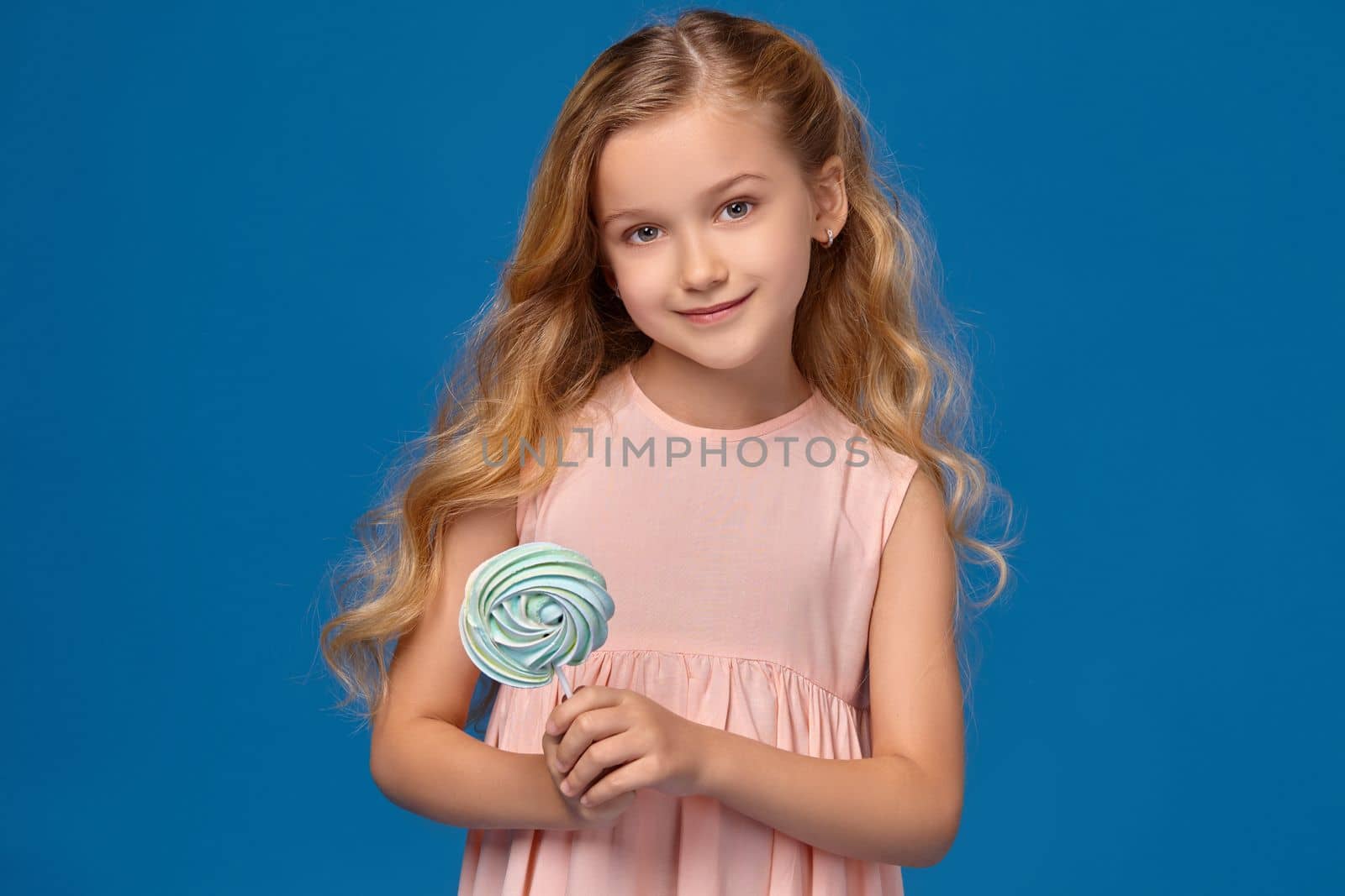 Nice little girl in a pink dress is holding a candy and smiling at the camera, on a blue background.