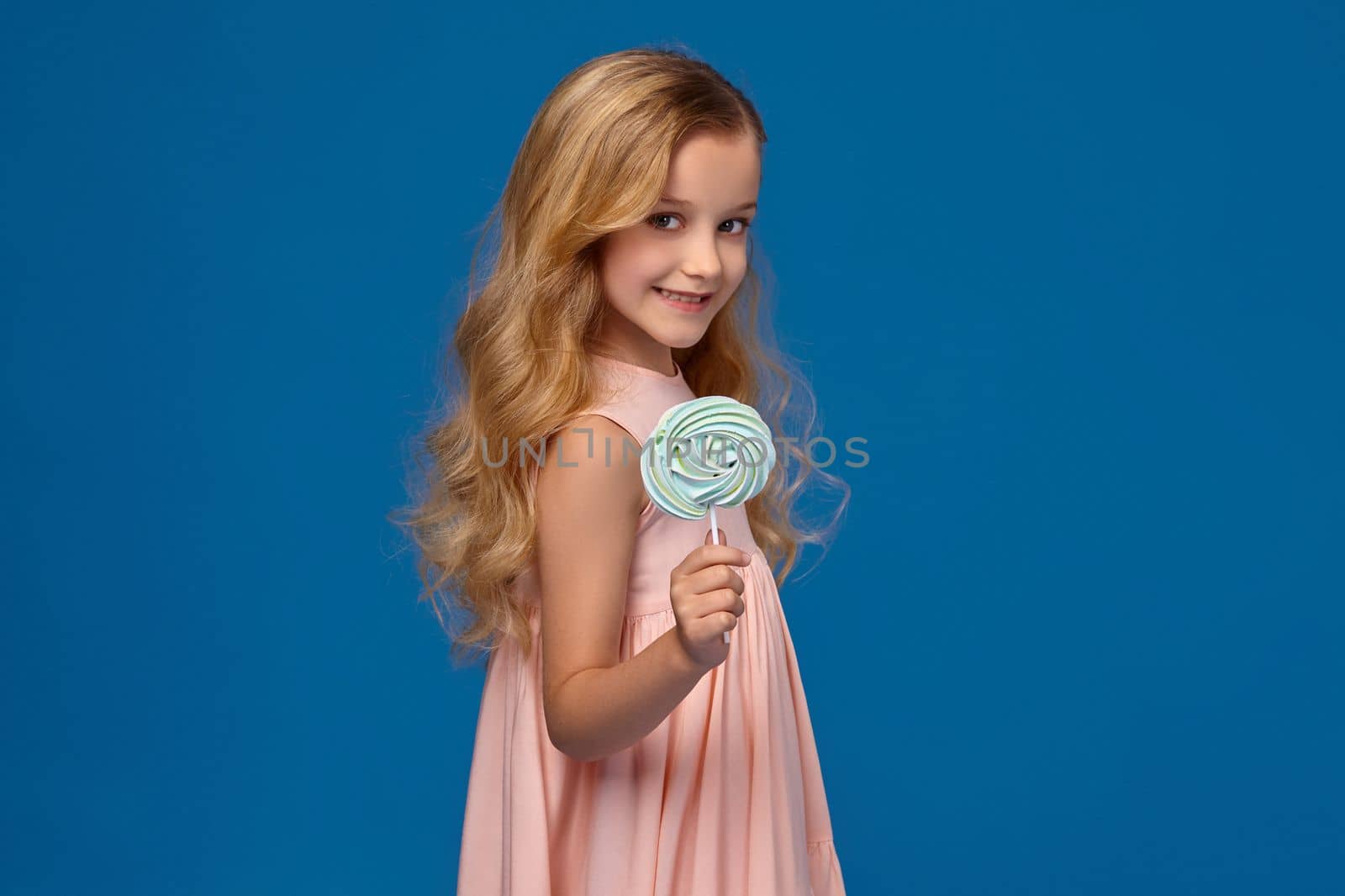 Little girl in a pink dress is holding a candy and showing it off at the camera, on a blue background.
