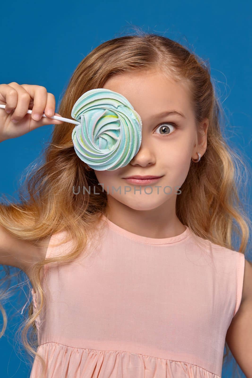 Little girl in a pink dress closes her eye with a candy, standing on a blue background.