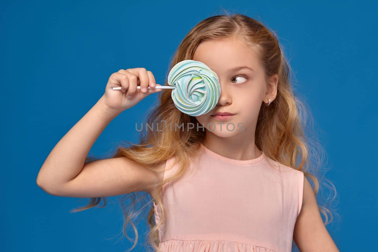 Pretty little girl in a pink dress closes her eye with a candy and looks away, standing on a blue background.
