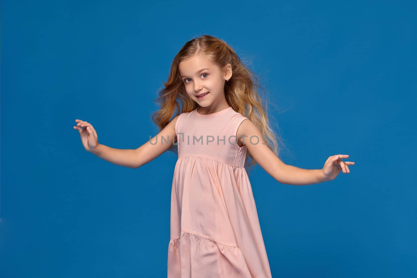 Fashionable little girl in a pink dress is dancing and looking happy, on a blue background.