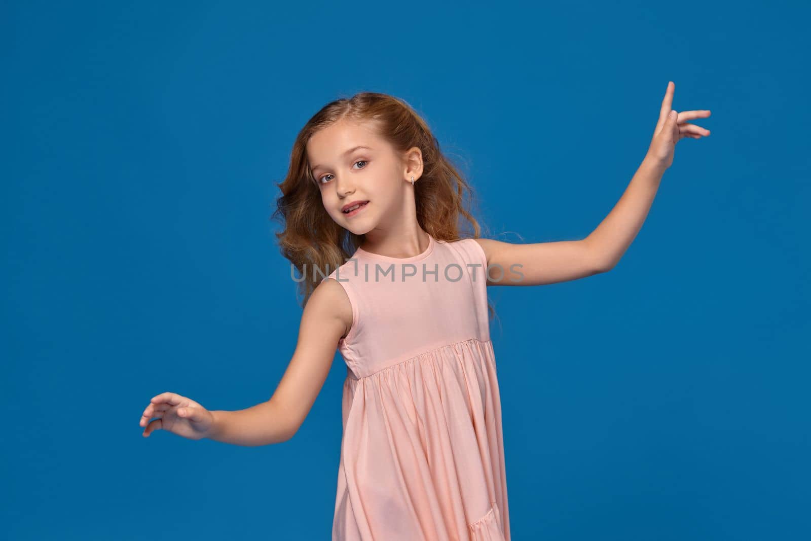 Pretty little girl in a pink dress, with a curly hair is dancing and looking happy, on a blue background.