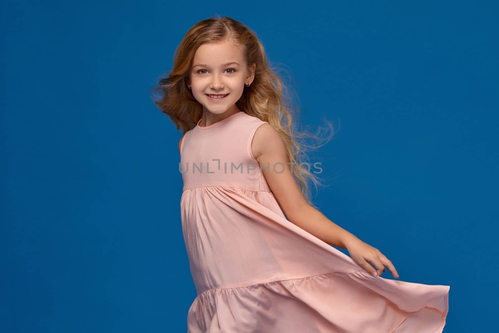 Beautiful little girl in a pink dress, with a curly hair is spinning around, on a blue background.