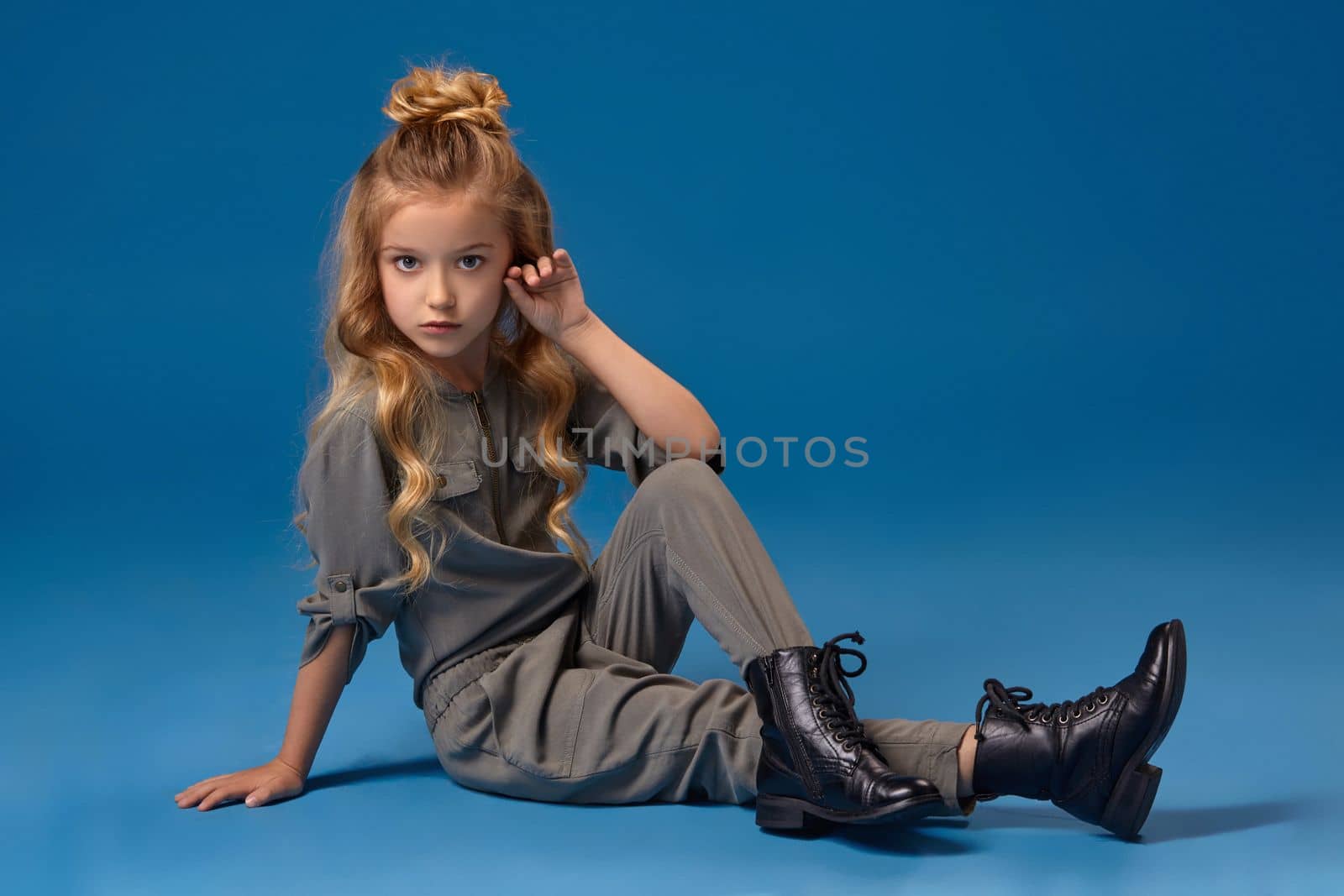 Little girl with a curly blond hair in a fashionable gray overall and black boots, set down and crossed her legs, on a blue background