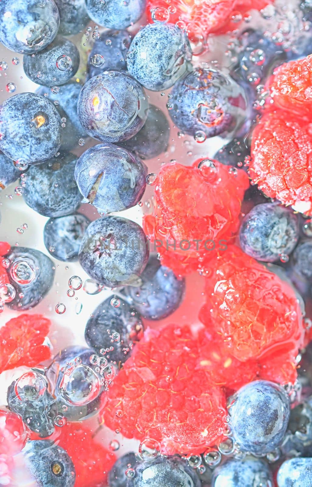 Blueberries and raspberries in liquid with bubbles. Colourful ripe bilberries and raspberries in water. Close-up of fresh berries in water background. Top view, flat design. Vertical macro image by roman_nerud