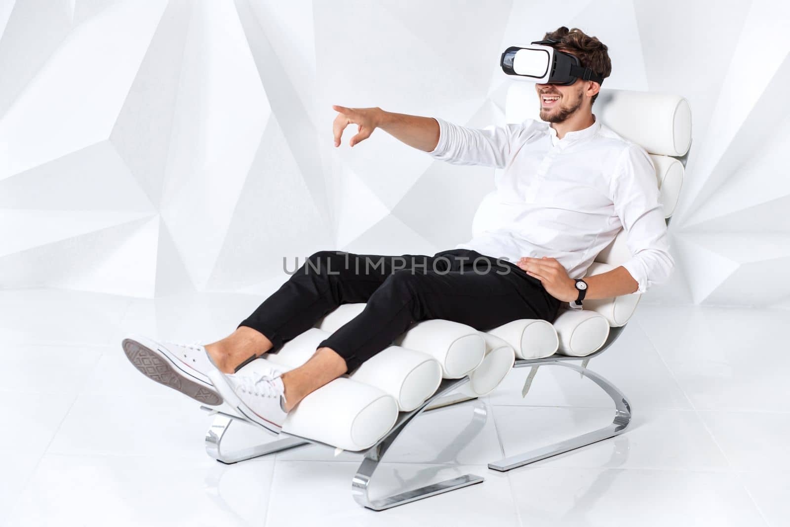 Excited young man is getting experience using VR-headset glasses of virtual reality gesticulating with his hands. A young man sits on a comfortable armchair in a room with white walls