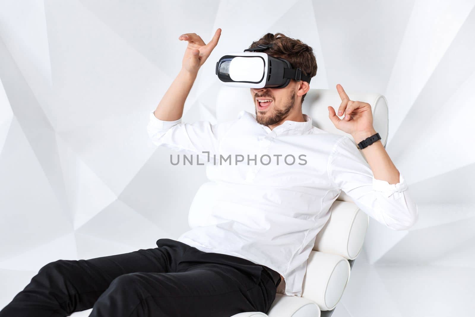 Excited young man is getting experience using VR-headset glasses of virtual reality gesticulating with his hands. A young man sits on a comfortable armchair in a room with white walls