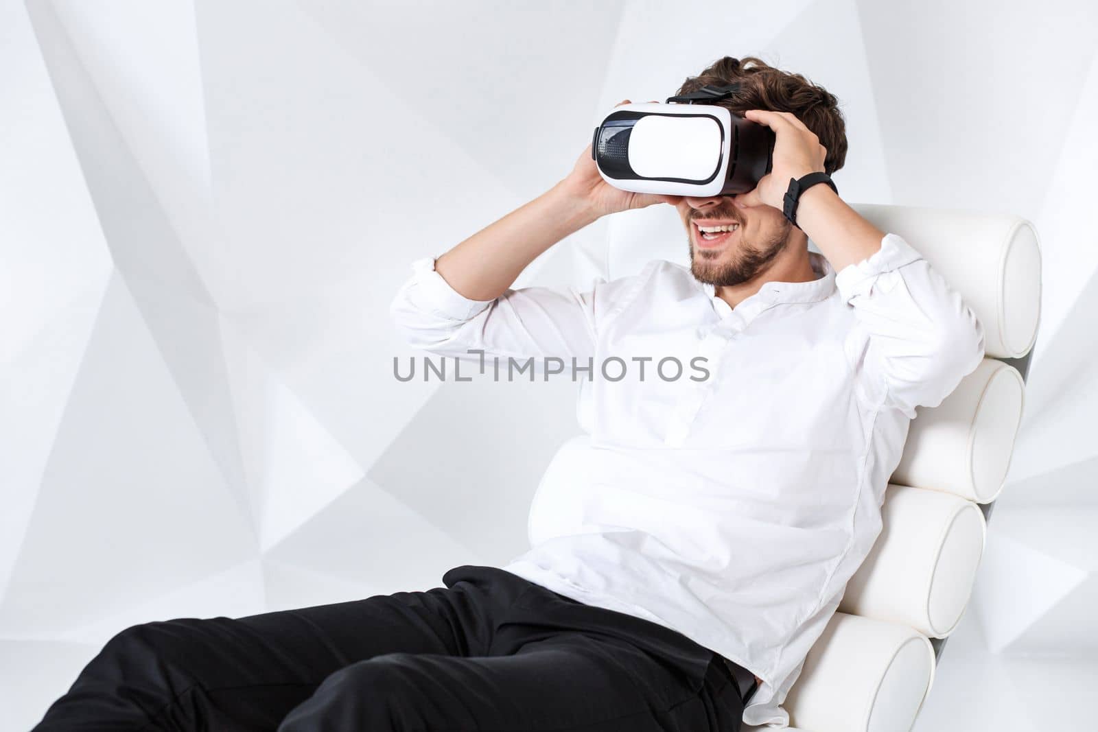Excited young man is getting experience using VR-headset glasses of virtual reality gesticulating with his hands. A young man sits on a comfortable armchair in a room with white walls