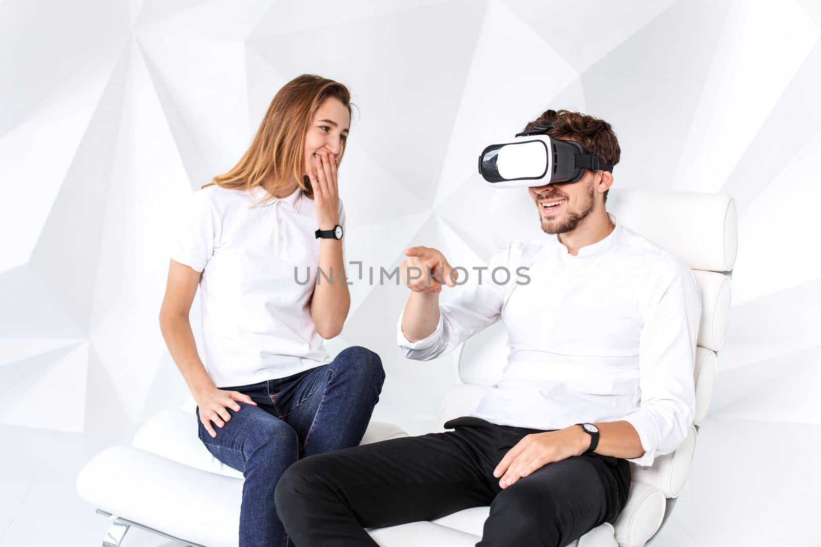 Couple having fun playing with virtual reality. A young man sits on a comfortable armchair in a room with white walls