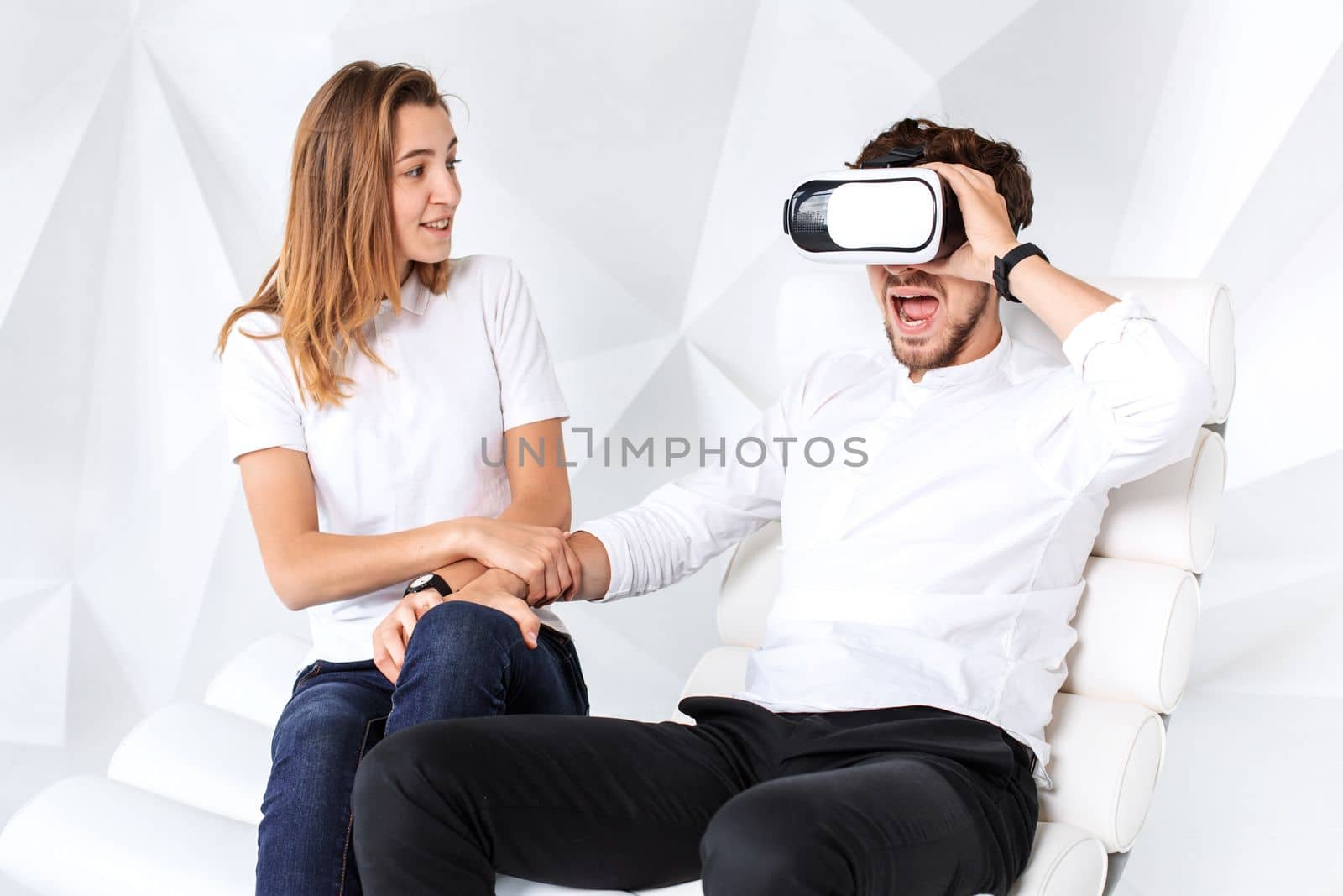 Couple having fun playing with virtual reality. A young man sits on a comfortable armchair in a room with white walls