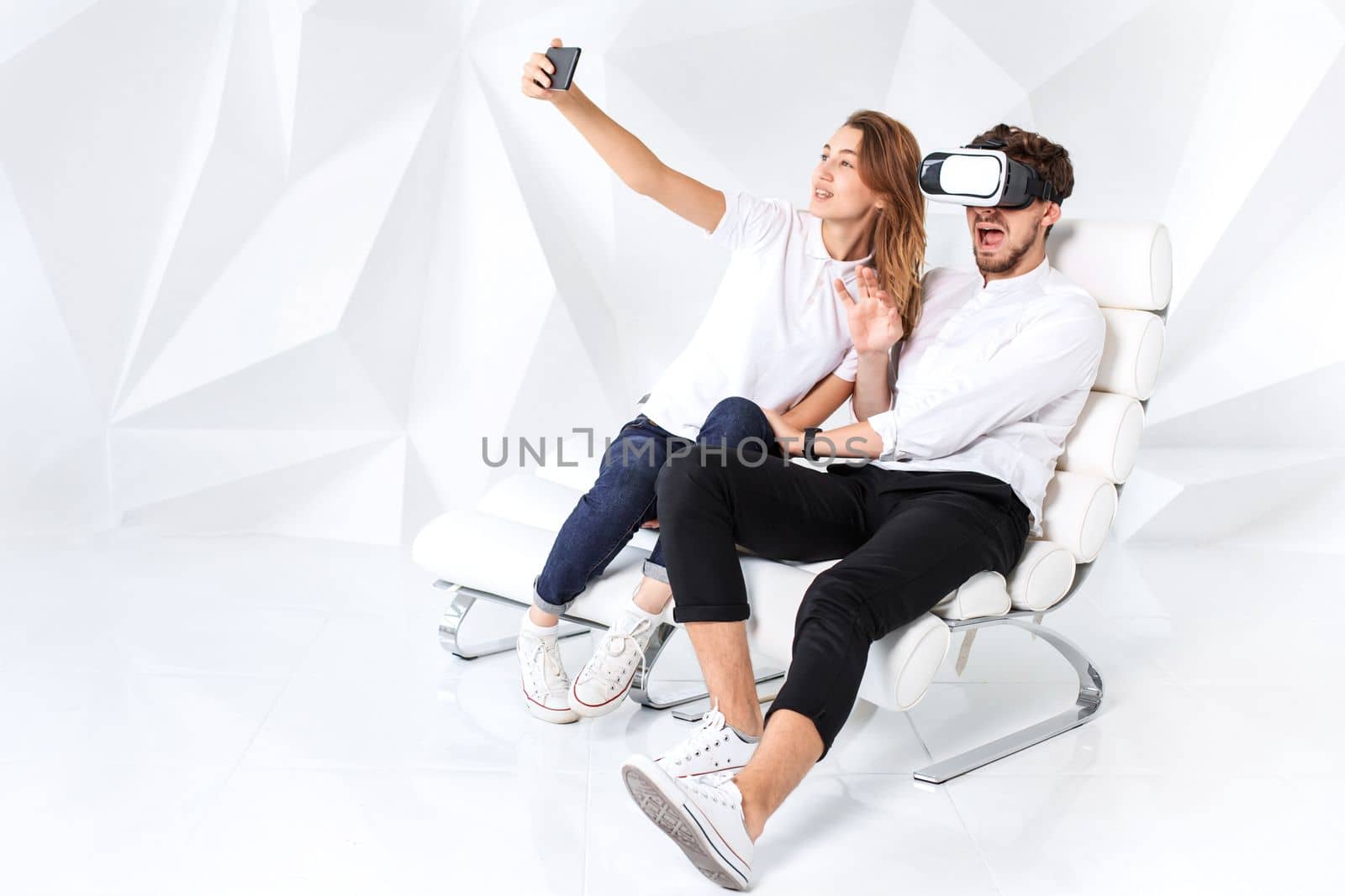 Couple having fun playing with virtual reality. A young man sits on a comfortable armchair in a room with white walls