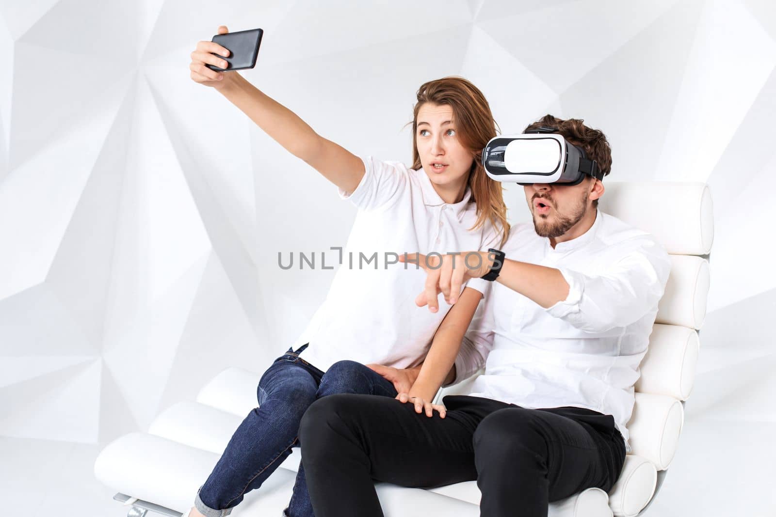 Couple having fun playing with virtual reality. A young man sits on a comfortable armchair in a room with white walls