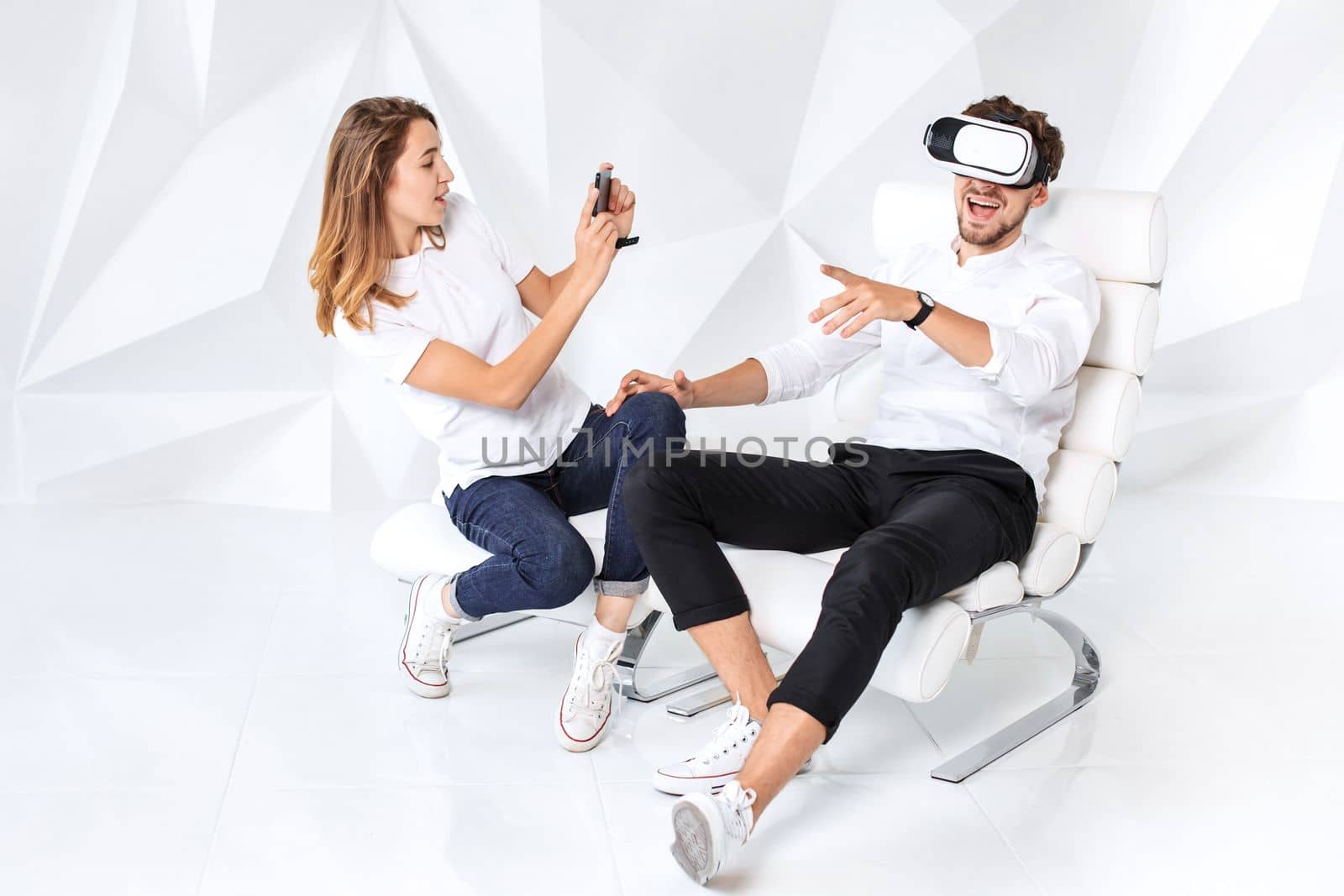 Couple having fun playing with virtual reality. A young man sits on a comfortable armchair in a room with white walls