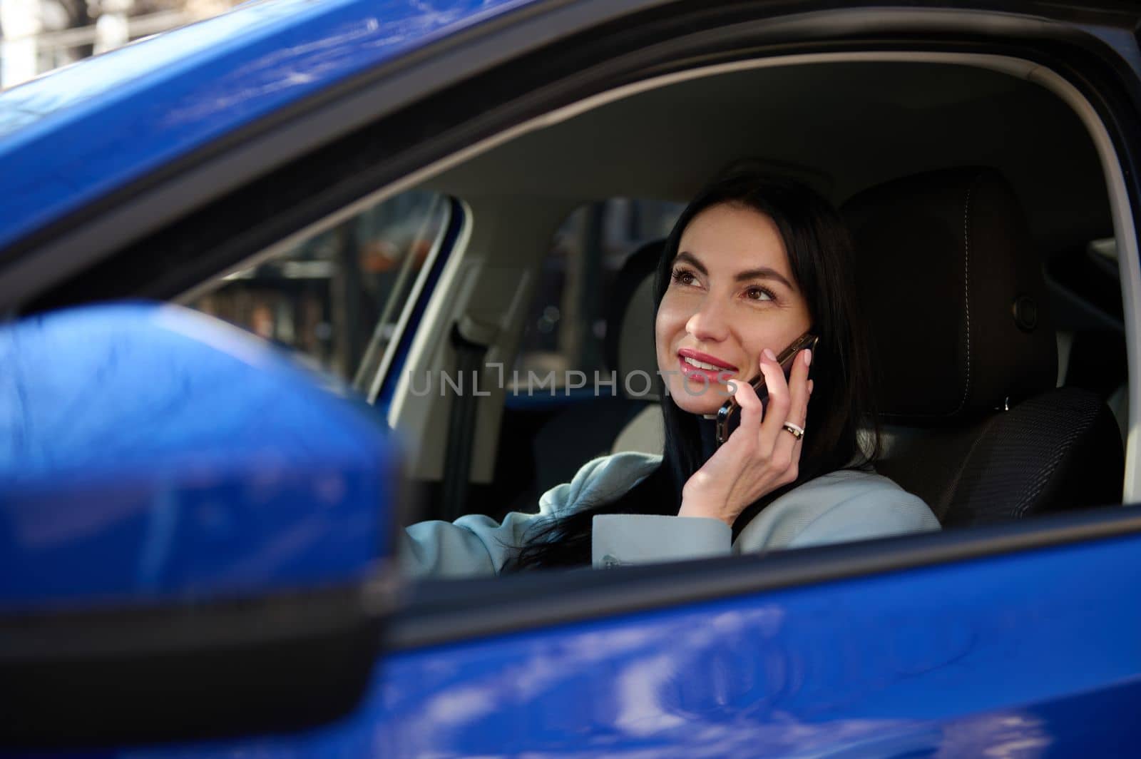 Close-up attractive stylish woman sitting in car on driver's seat, dressed in coat winter style, using mobile phone, talking with clients, plans business meeting. Successful businesswoman driving car