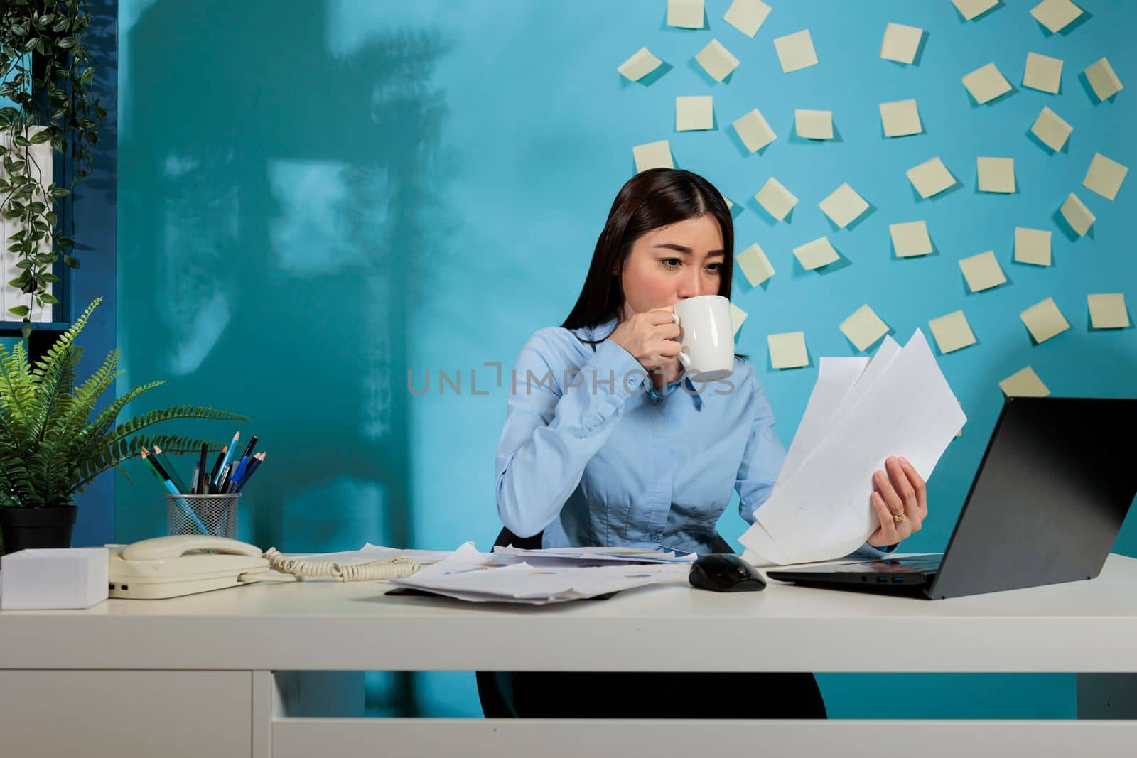 Corporate female auditor sitting at office desk analyzing financial reports of startup company. Professional woman drinking cup of coffee while comparing digital information and printed paperwork.