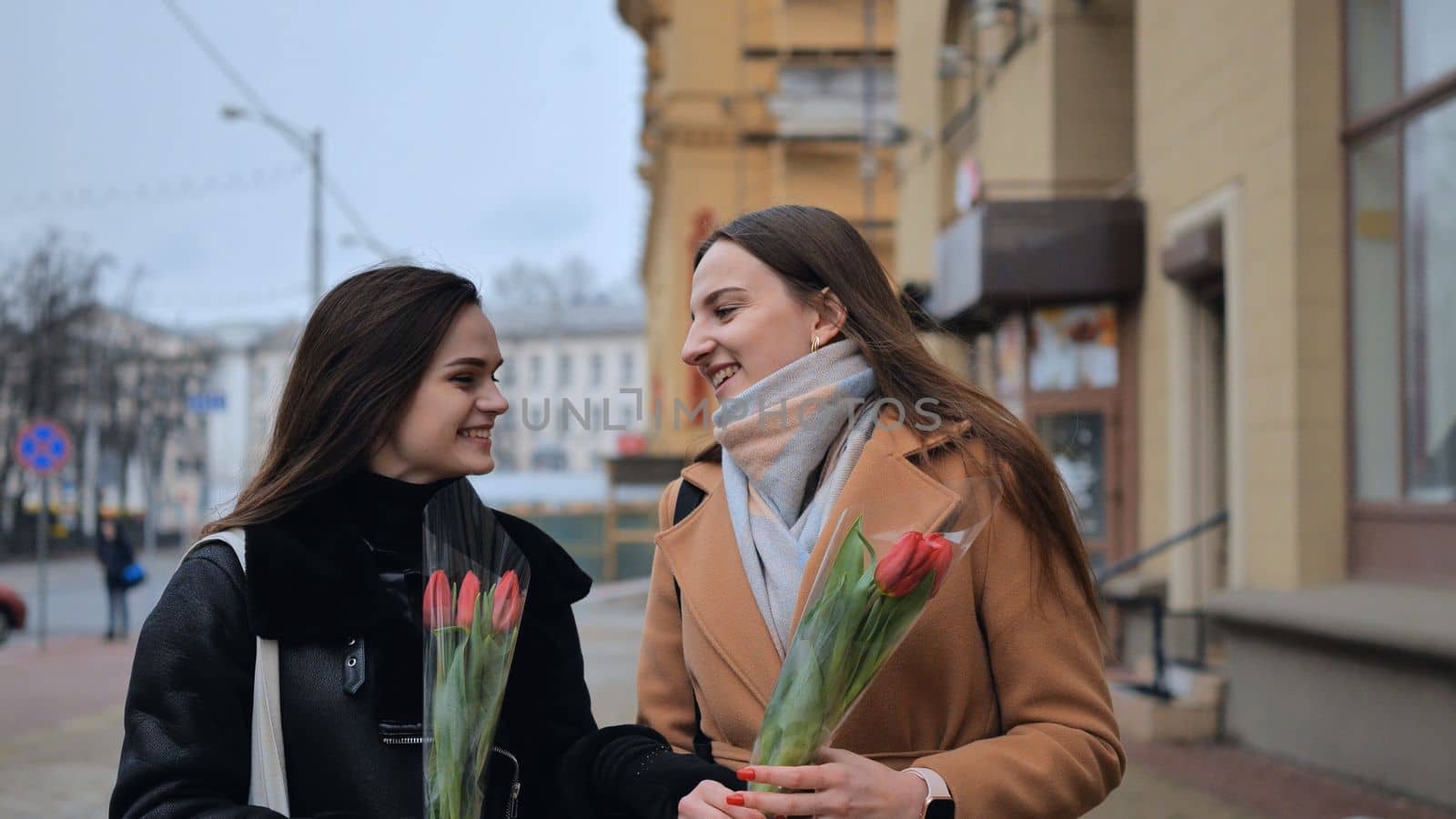 Two joyful friends in a jacket and coat are walking around the city with tulips