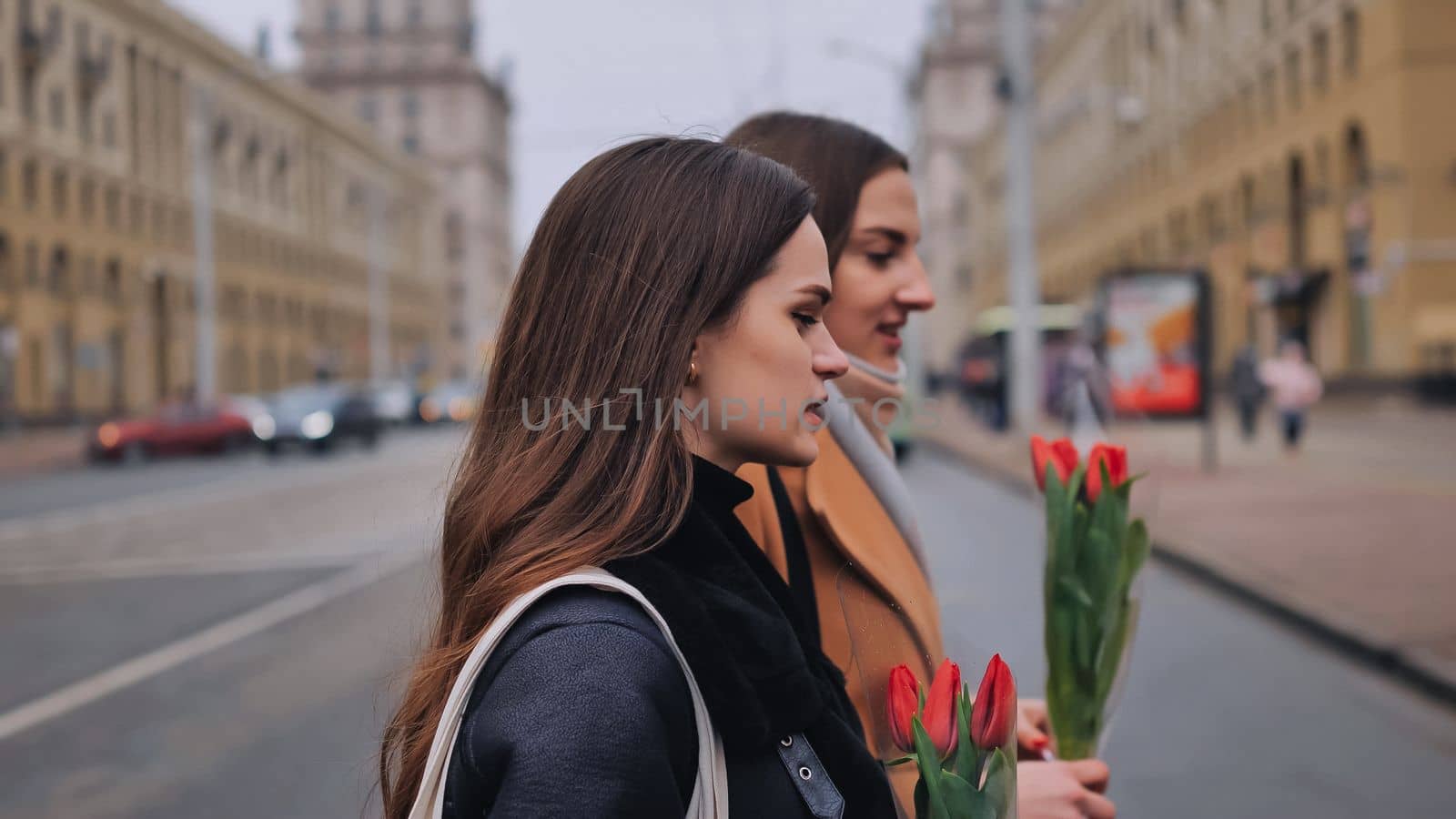 Two joyful friends in a jacket and coat are walking around the city with tulips
