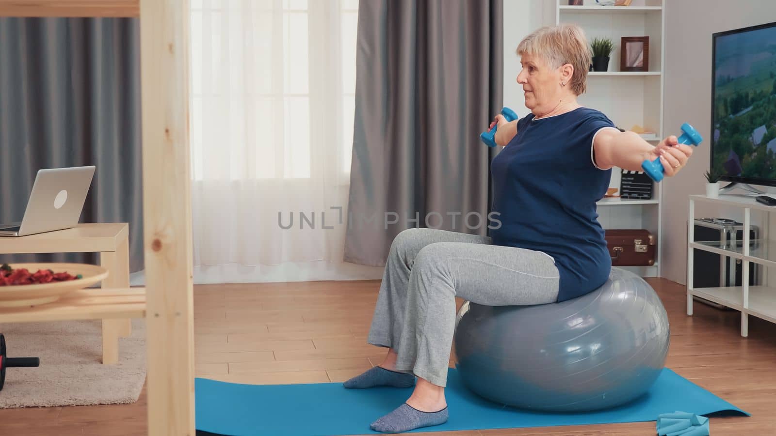 Grandmother lifting weights sitting on balance ball in living room. Old person training at home sport healthy lifestyle, elderly fitness exercise workout in apartment, activity and healthcare