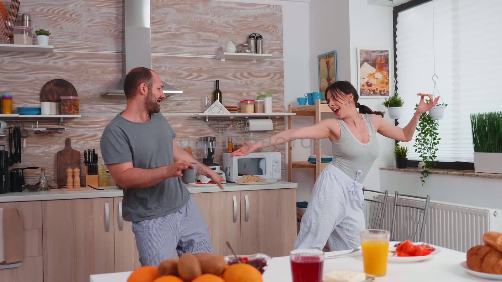 Married couple having fun dancing in kitchen during breakfast. Carefree husband and wife laughing, singing, dancing listening musing, living happy and worry free. Positive people.