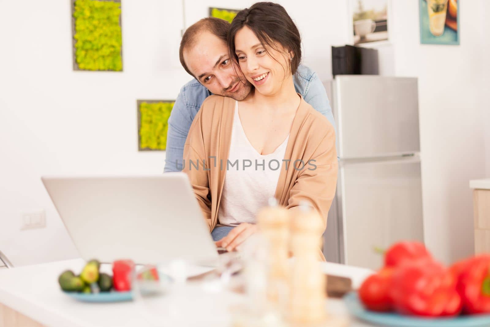 Smiling couple using laptop in kitchen with healthy vegetables on the table. Happy loving cheerful romantic in love couple at home using modern wifi wireless internet technology