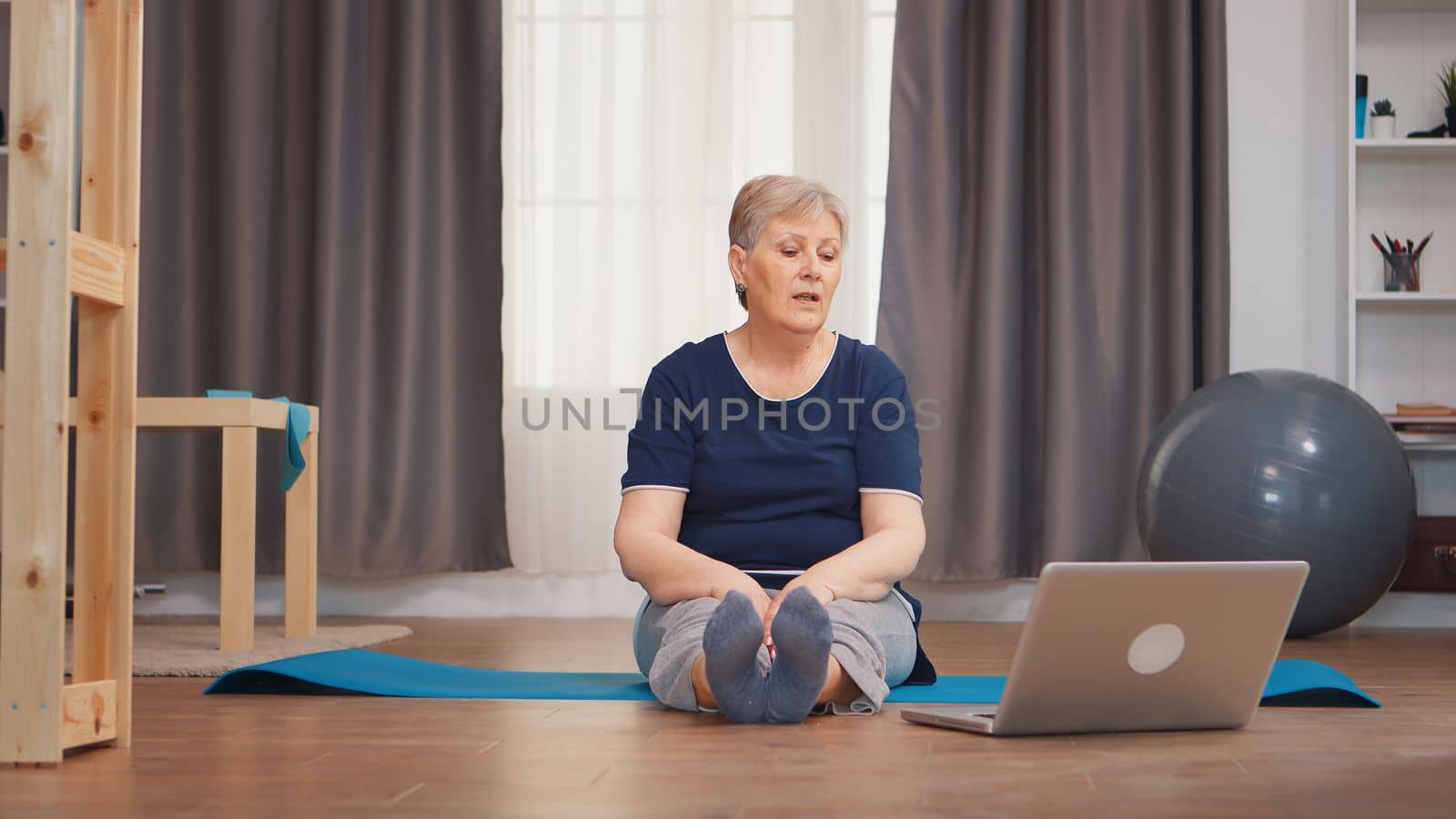 Senior woman sitting on yoga mat stretching boyd sitting on mat. Online learning and study, Active healthy lifestyle sporty old person training workout home wellness and indoor exercising