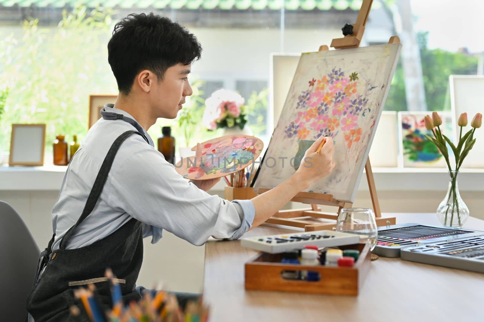 Satisfied man painting on easel with watercolor in comfortable art workshop. Leisure activity and creative hobby concept.