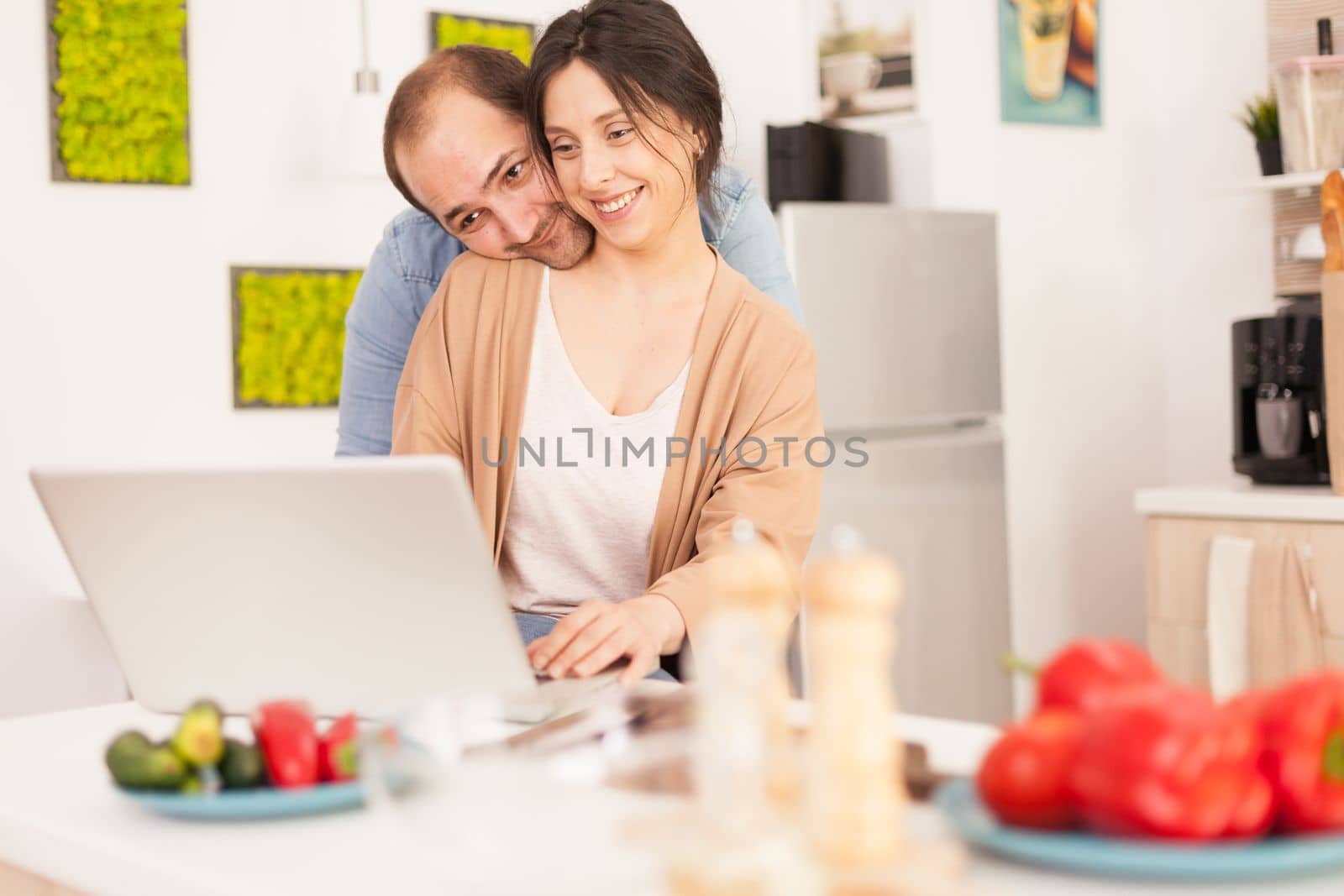 Boyfriend hugs girlfriend in kitchen and she is using laptop. Happy wife. Fresh vegetables. Happy loving cheerful romantic in love couple at home using modern wifi wireless internet technology