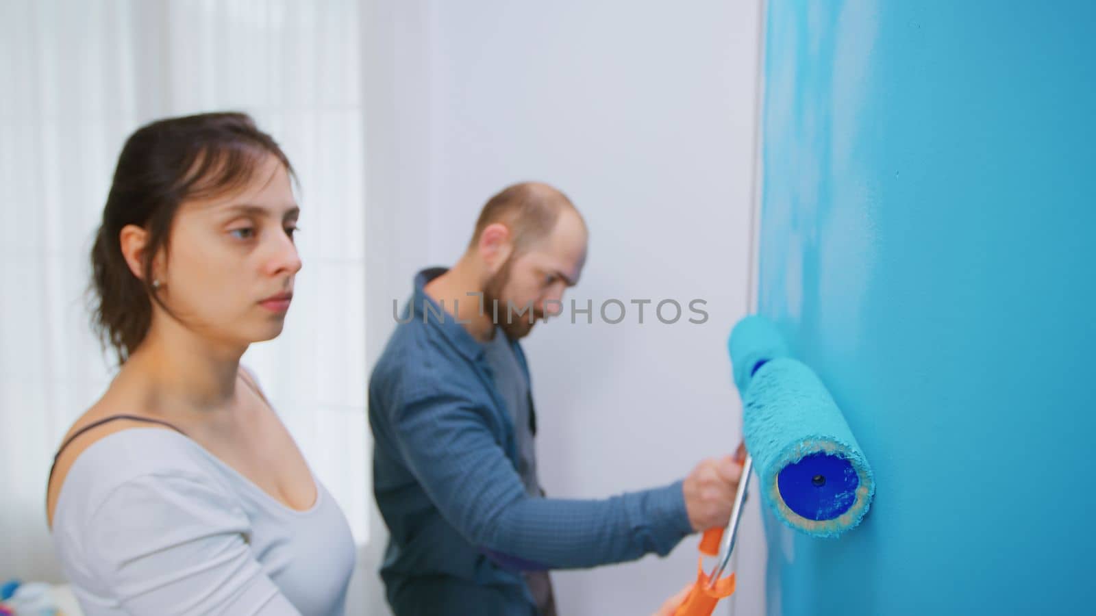 Woman painting wall with roller brush while redecorating apartment living room. Couple redecorating together. Apartment redecoration and home construction while renovating and improving. Repair and decorating.